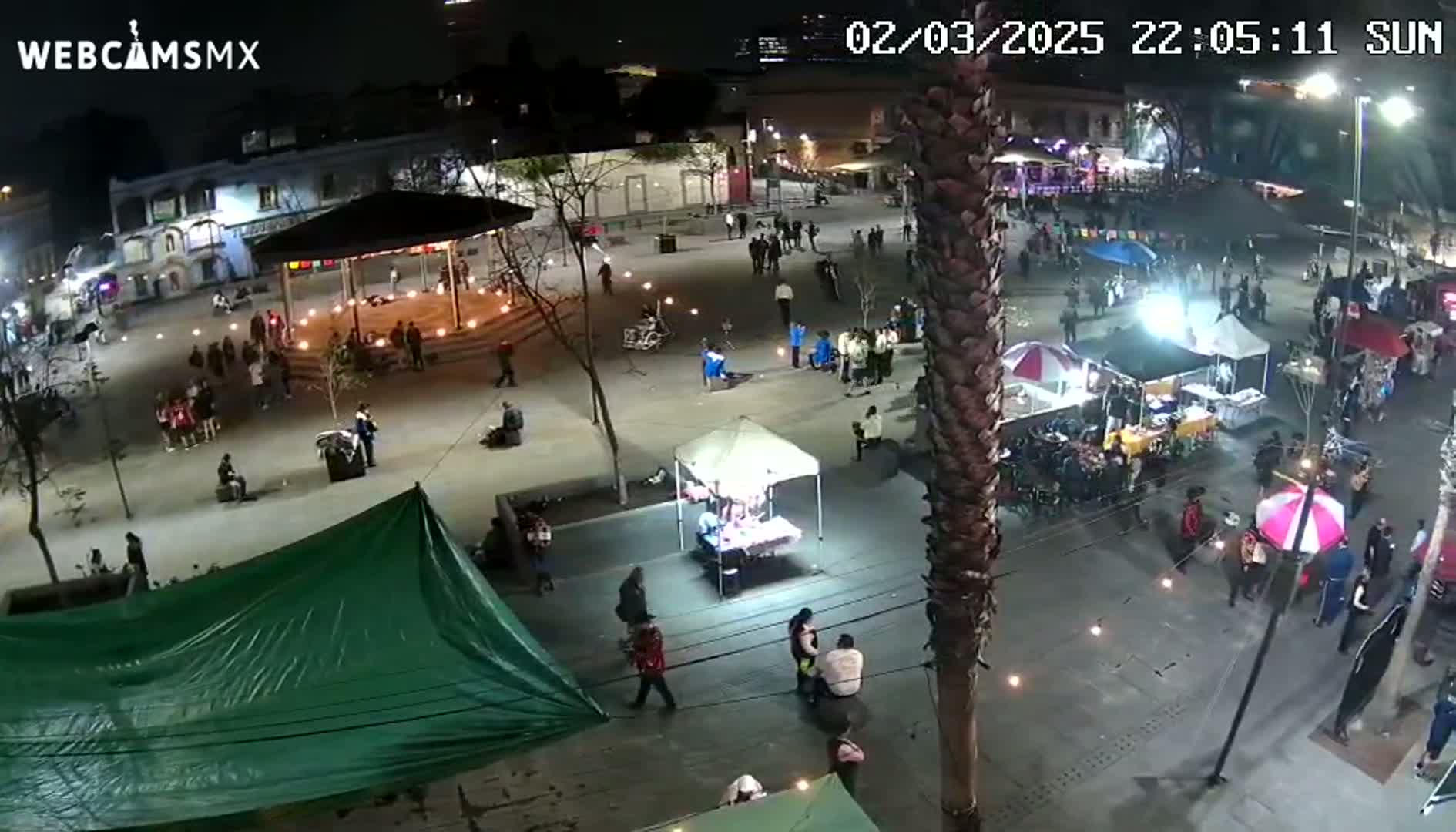 A nighttime, outdoor plaza scene shows numerous people gathered around various food stalls and vendors under ambient lighting.