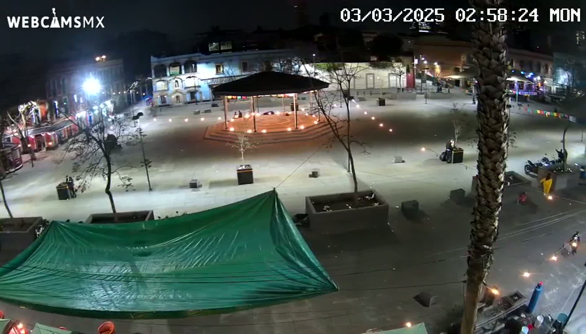 A nighttime view of a town square with a gazebo, illuminated by lights, surrounded by buildings and a large green tarp.