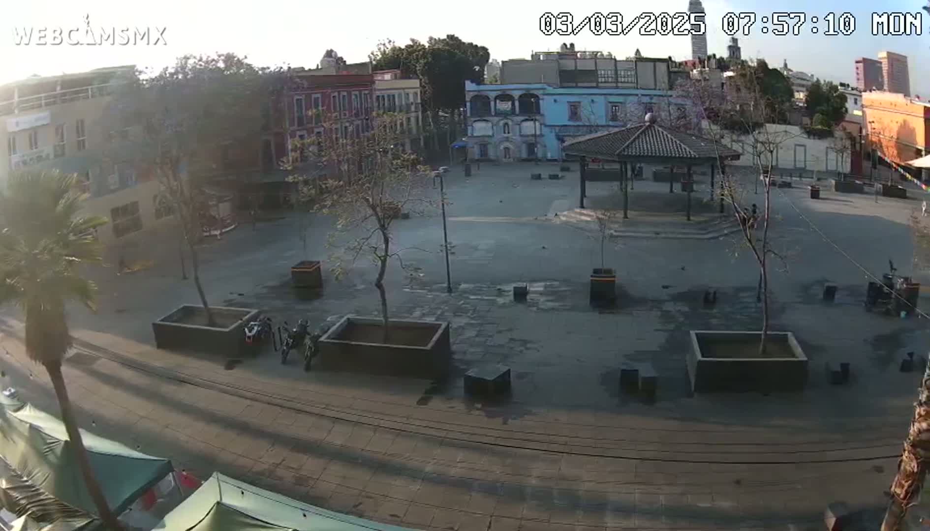 A mostly empty plaza, under a partly sunny sky, is surrounded by buildings and a few parked motorcycles.