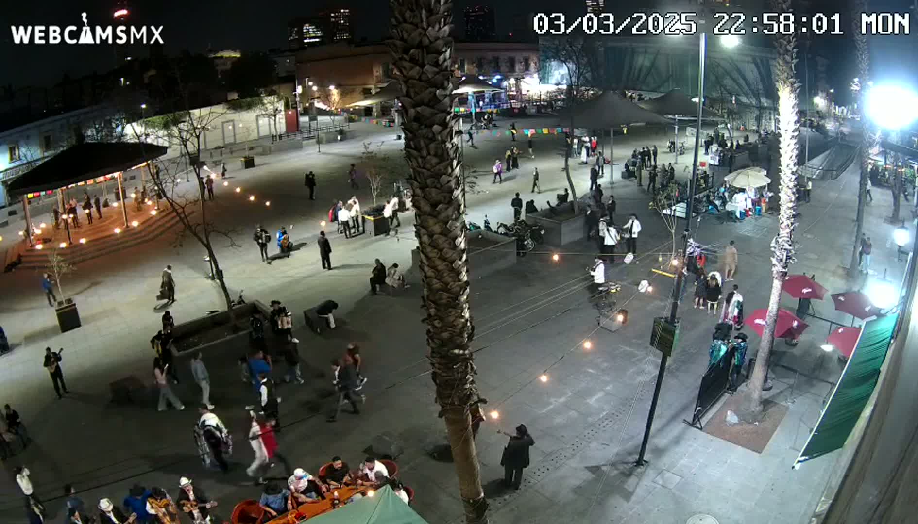A nighttime, outdoor plaza scene shows numerous people milling about, with several small groups of musicians performing, and some people sitting at outdoor tables under umbrellas.