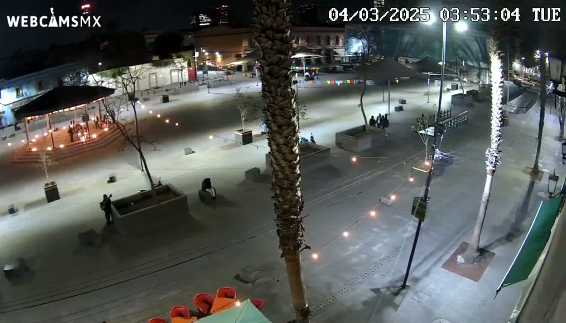 A nighttime view of a plaza with a few people scattered throughout, illuminated by string lights and building lights.