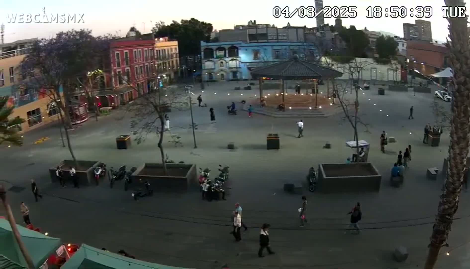 A high-angle, dusk-time view shows a town square with people walking around, some motorbikes parked, and a gazebo in the center.