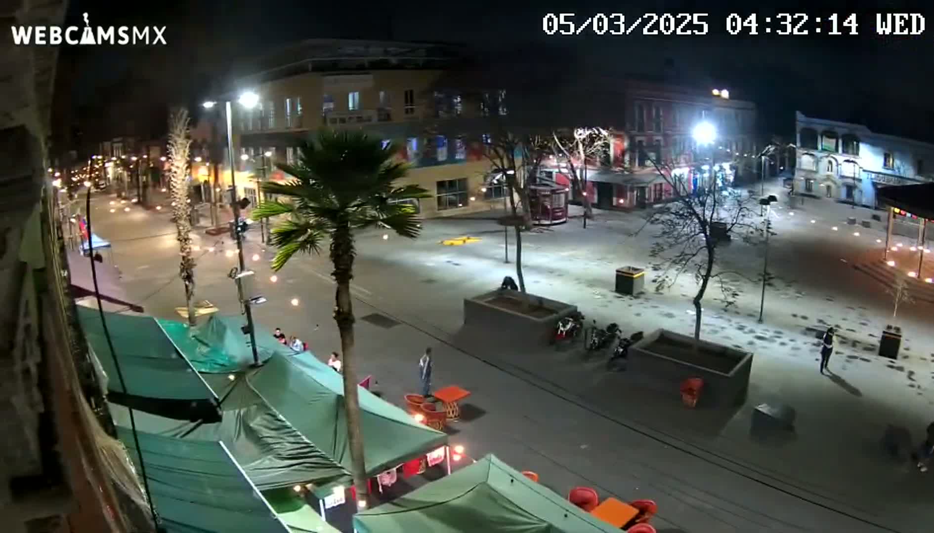 A nighttime, brightly-lit city square with a few pedestrians, several parked motorcycles, and numerous market stalls under green canopies.