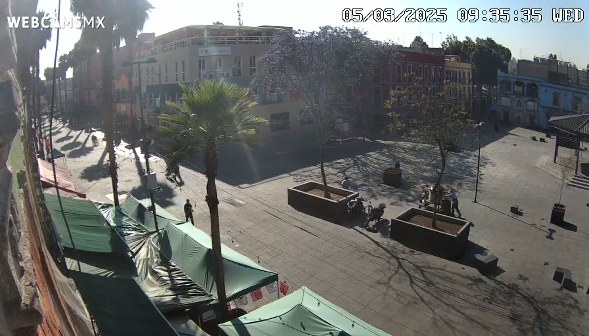 A sunny day reveals a town square with several people walking, some vendors with green canopies, and motorcyclists in a plaza surrounded by buildings and palm trees.