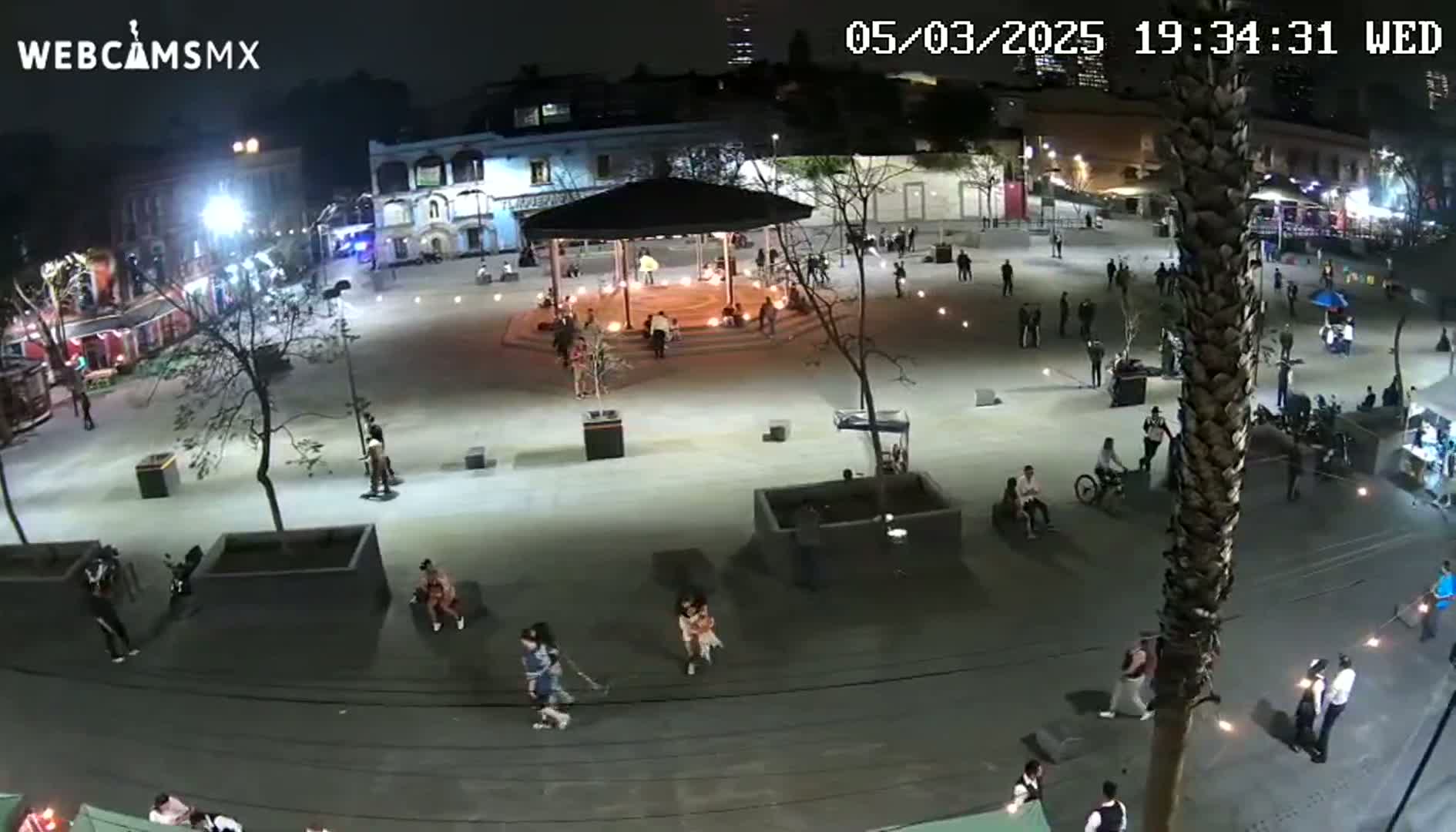 A nighttime, high-angle view of a plaza with people gathered around a gazebo, illuminated by artificial lights.