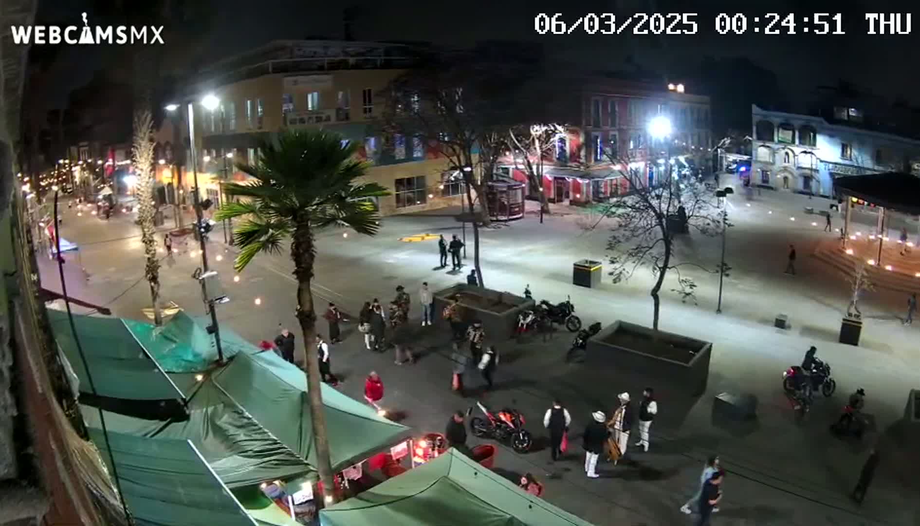 A nighttime view of a town square with people gathered around market stalls, motorcycles parked nearby, and palm trees lining the streets under bright lights.