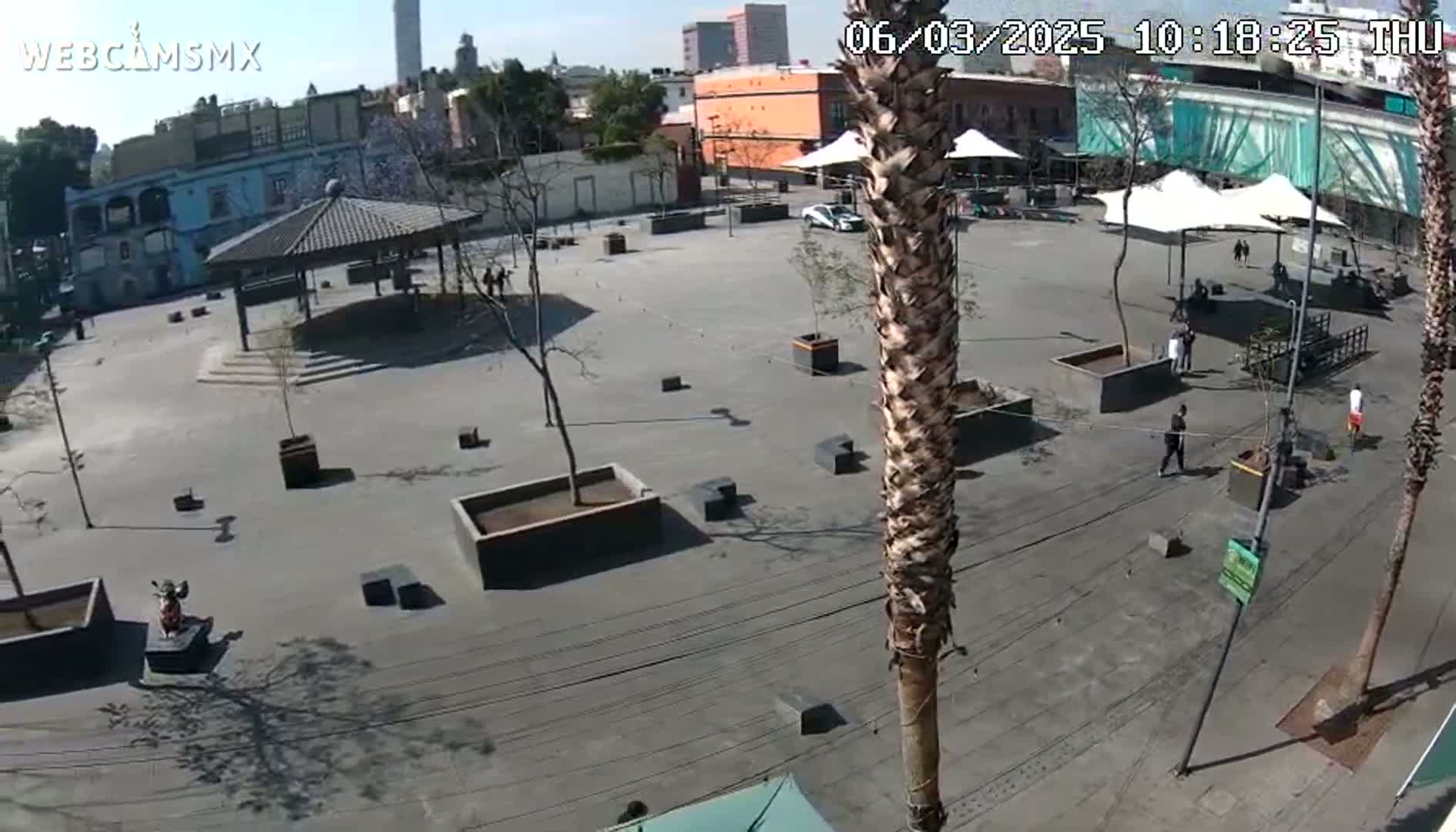 A mostly empty plaza with a gazebo, several palm trees, and some people is shown on a sunny day.