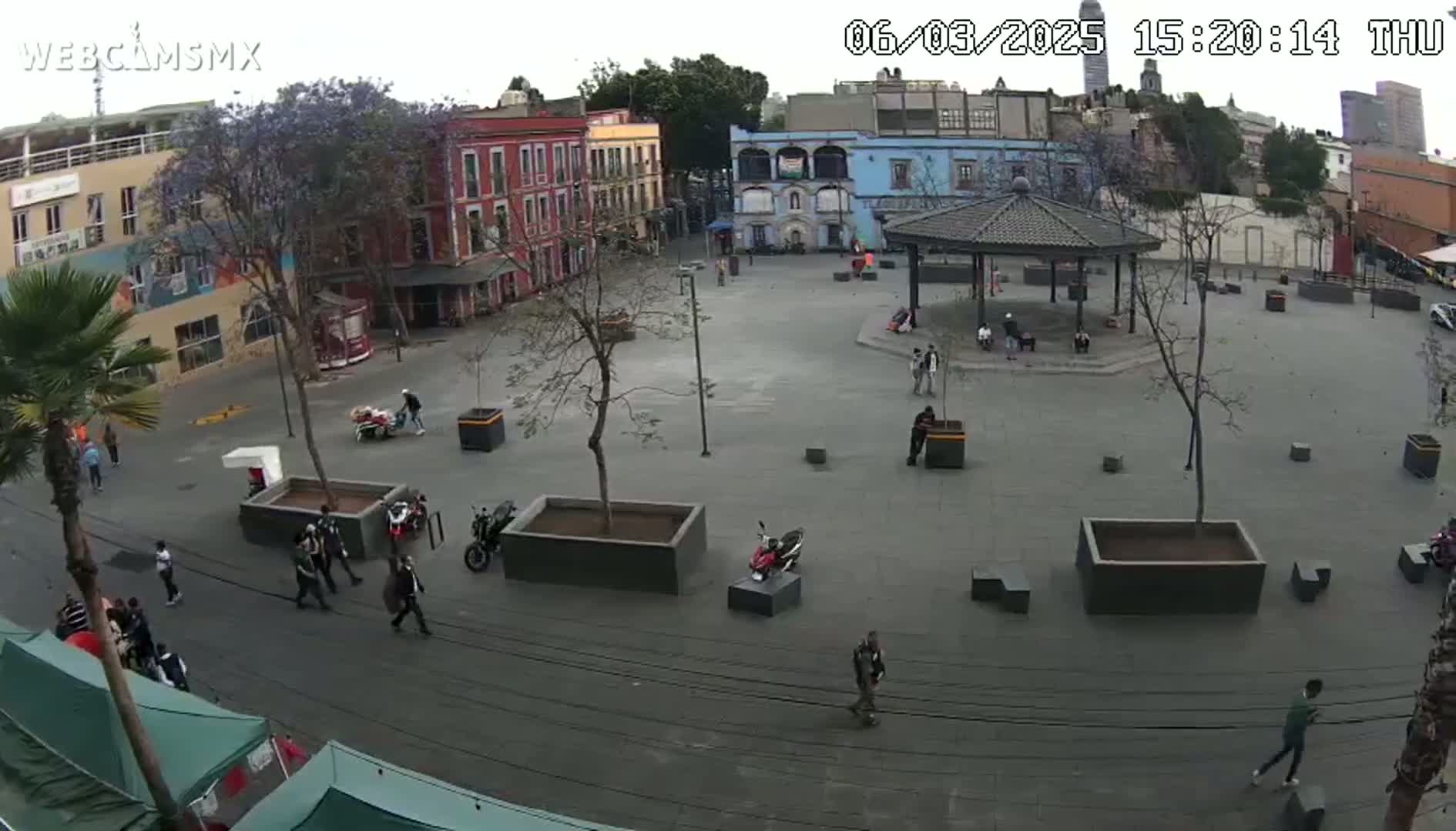 A mostly empty town square with several buildings surrounding it, a gazebo in the center, and a few people walking around under an overcast sky.