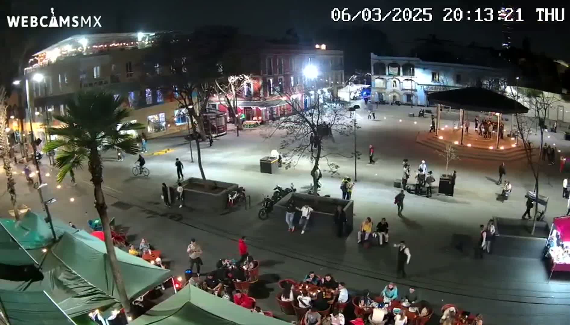 A nighttime aerial view of a town square, illuminated by streetlights, shows people gathered in small groups around tables and walking on paved pathways under a clear sky.