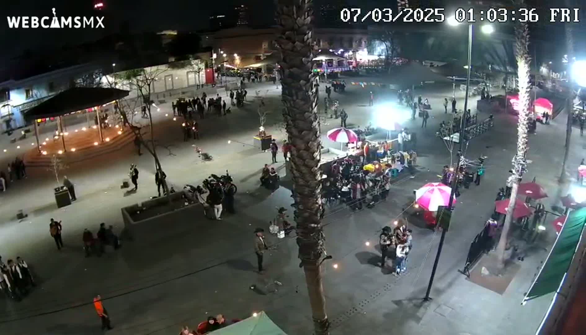 A nighttime aerial view of a town square filled with people, food vendors, and illuminated structures under a clear sky.