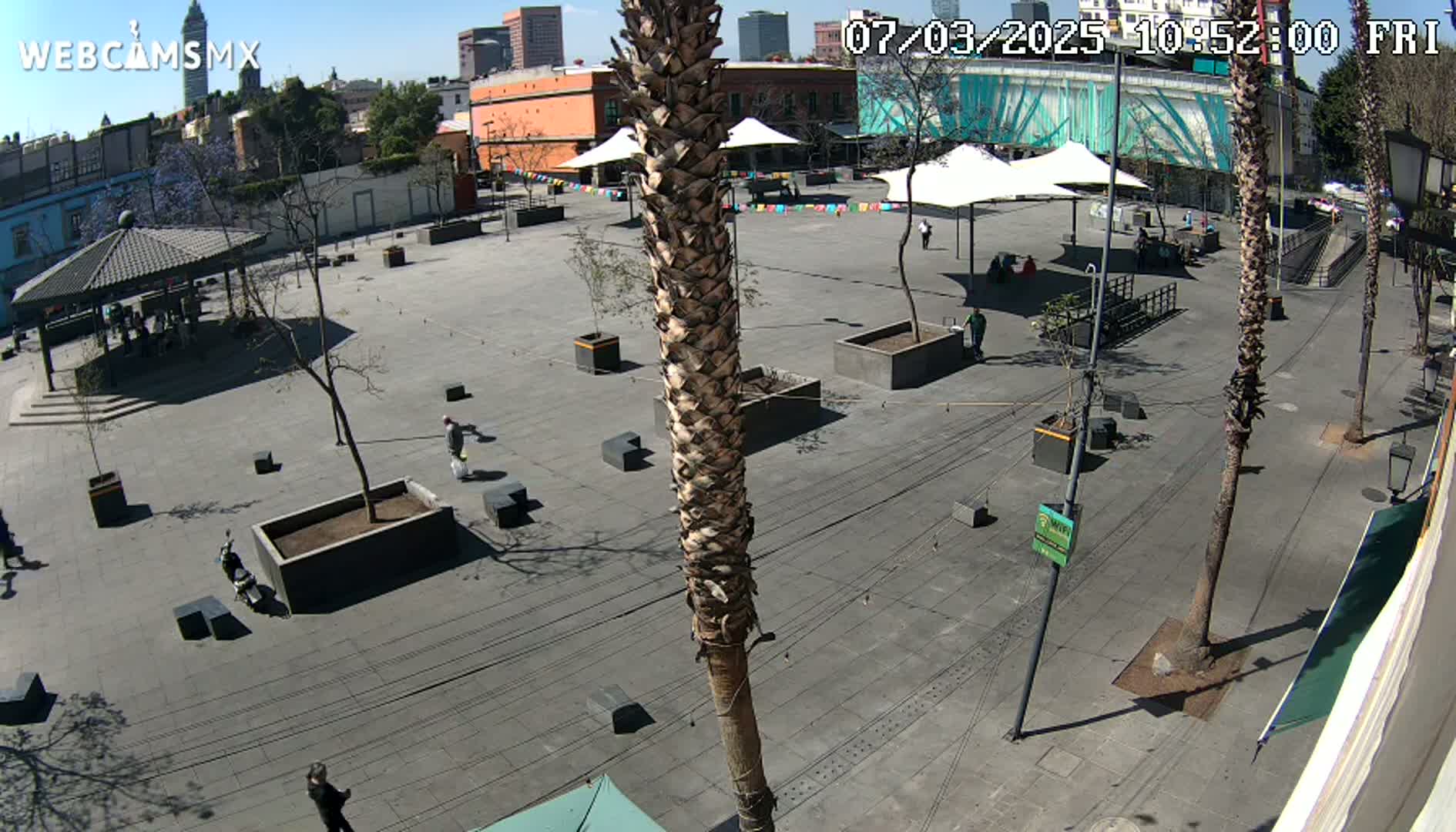 A sunny day in a plaza shows several people walking around, some sitting under shade umbrellas, with buildings and palm trees in the background.