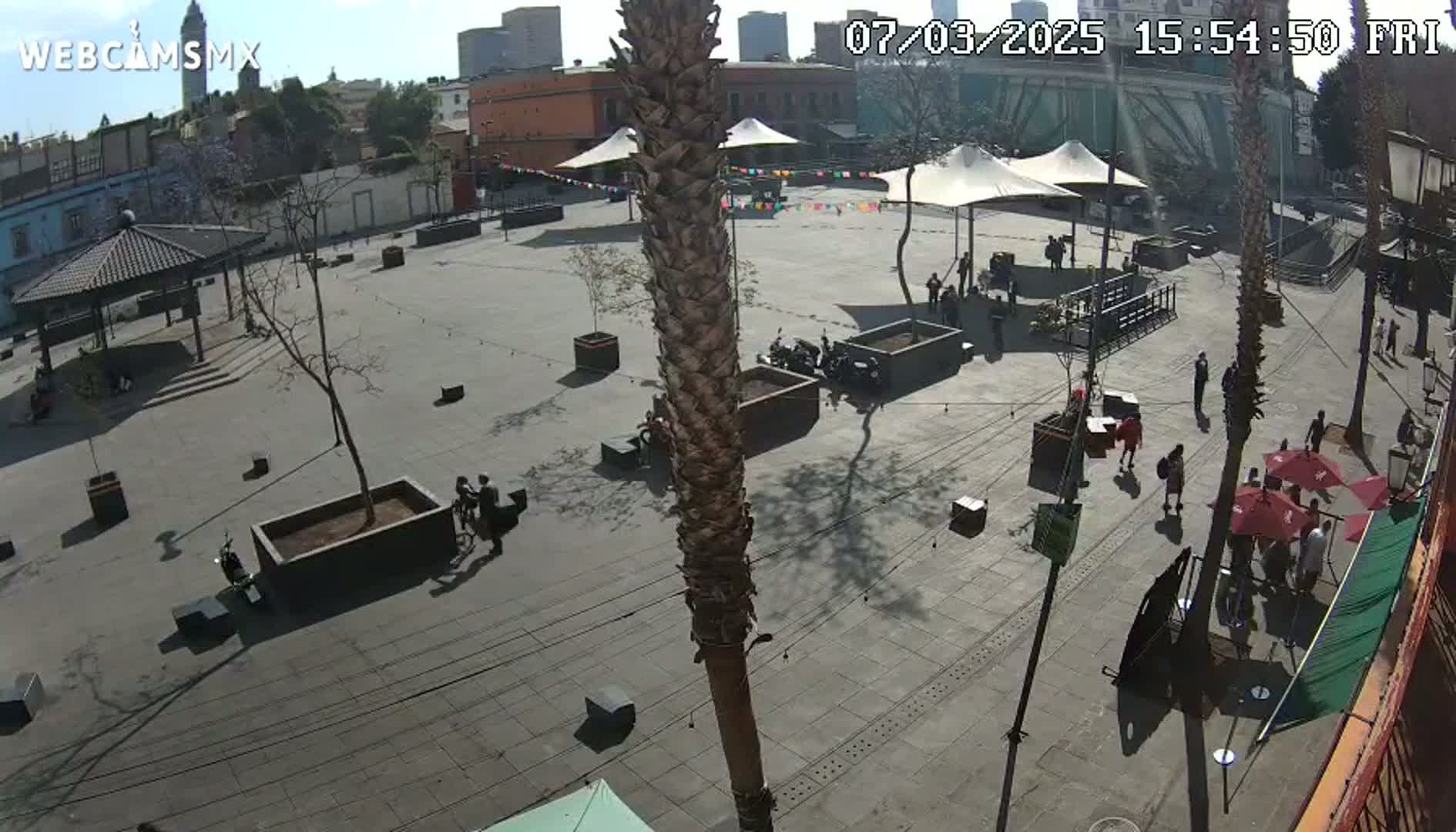 A sunny outdoor plaza with several people, some sitting at tables under red umbrellas, and others walking around, near buildings and palm trees.