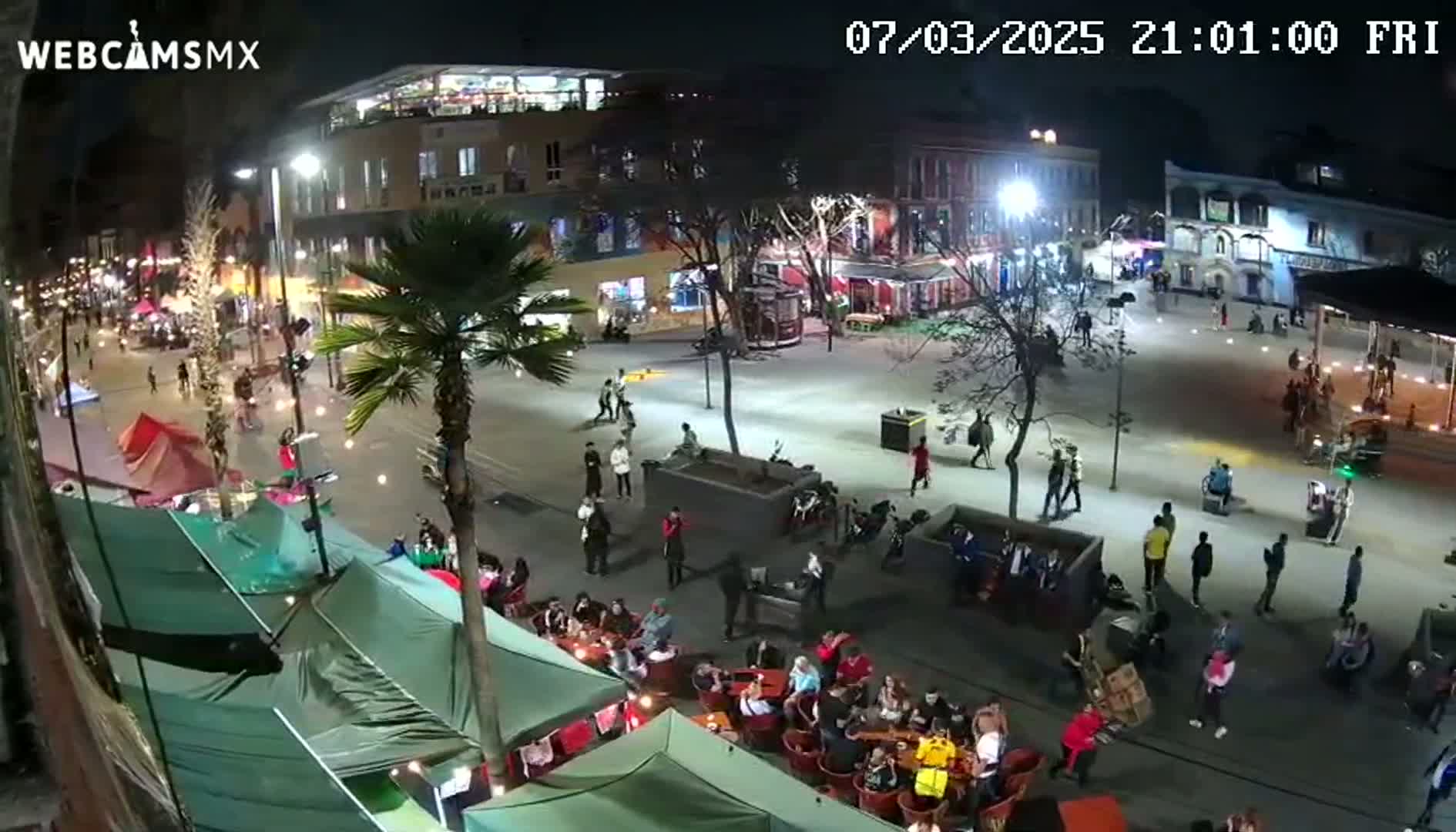 A nighttime, brightly lit plaza scene shows people walking around, sitting at outdoor cafes under green tents, and parked motorcycles, with palm trees and buildings lining the area.