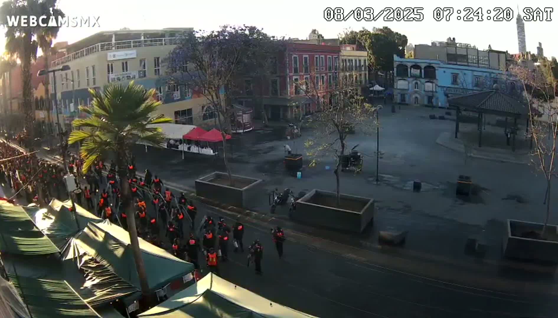 A large group of people in dark clothing walks down a street past a plaza with several buildings, a gazebo, and some parked motorcycles, under a partly sunny sky.