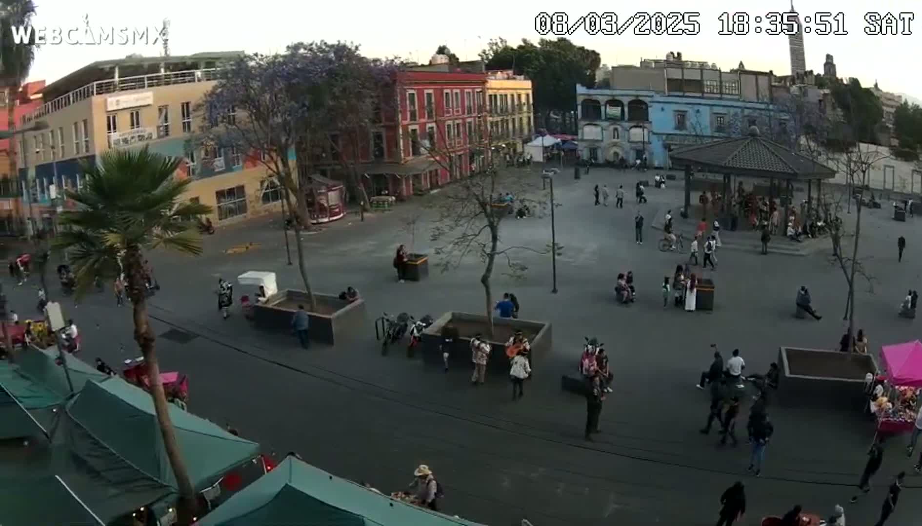 An overcast day reveals a town square with people walking and gathering around a gazebo, surrounded by buildings and street vendors.