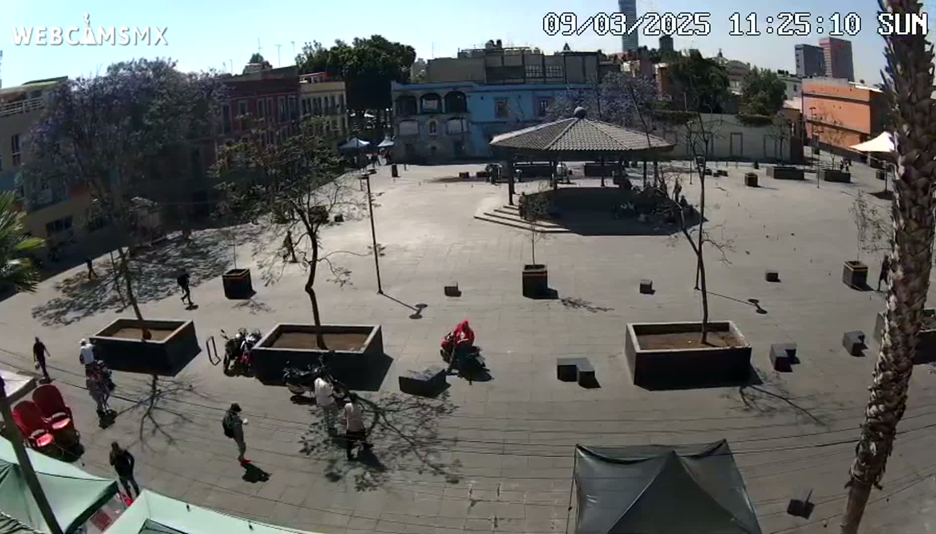 A mostly empty plaza with several buildings surrounding it on a sunny day, a few people are scattered throughout the area.