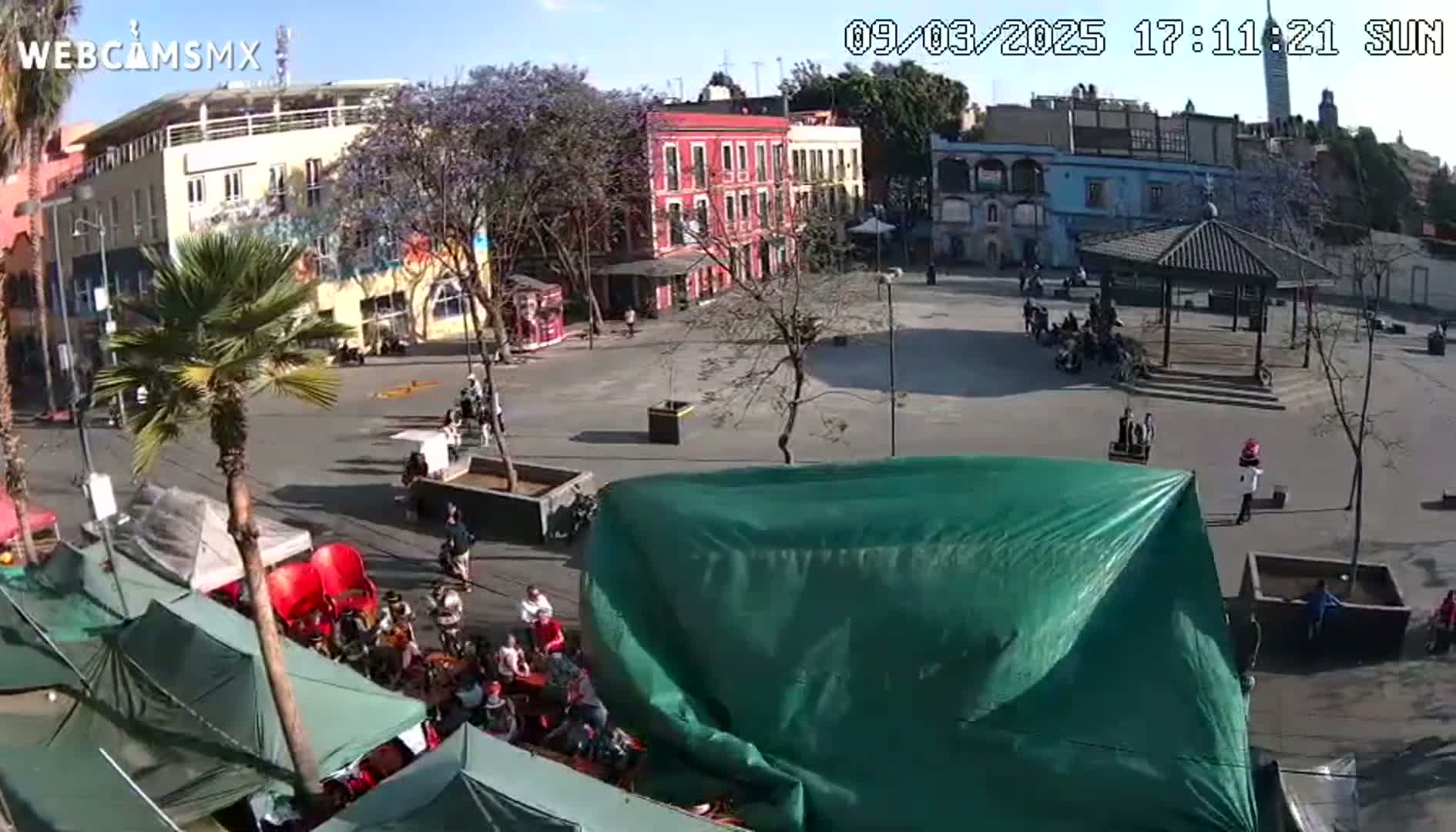 A sunny plaza scene shows several buildings, a gazebo, people walking and sitting, and numerous tents and canopies, with a large green tarp covering something in the foreground.