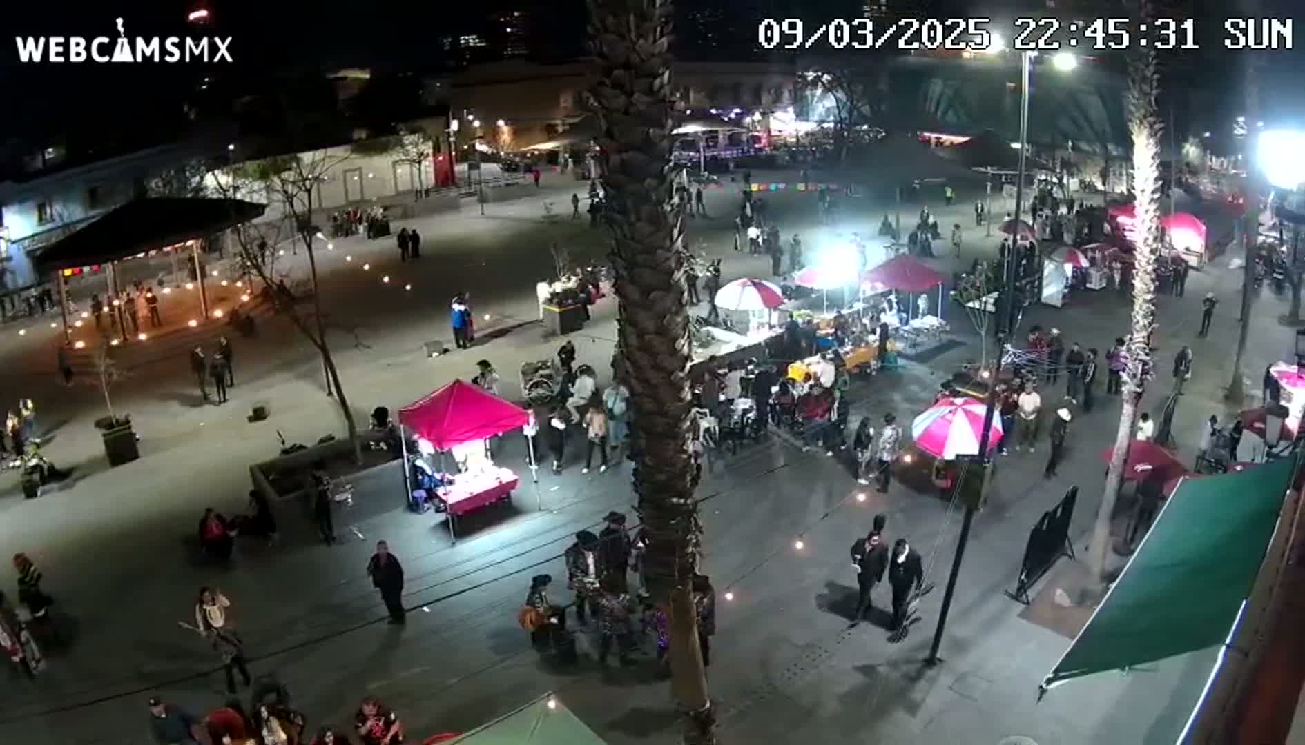 A nighttime aerial view shows a town square filled with people, food vendors, and small market stalls under the illumination of streetlights.