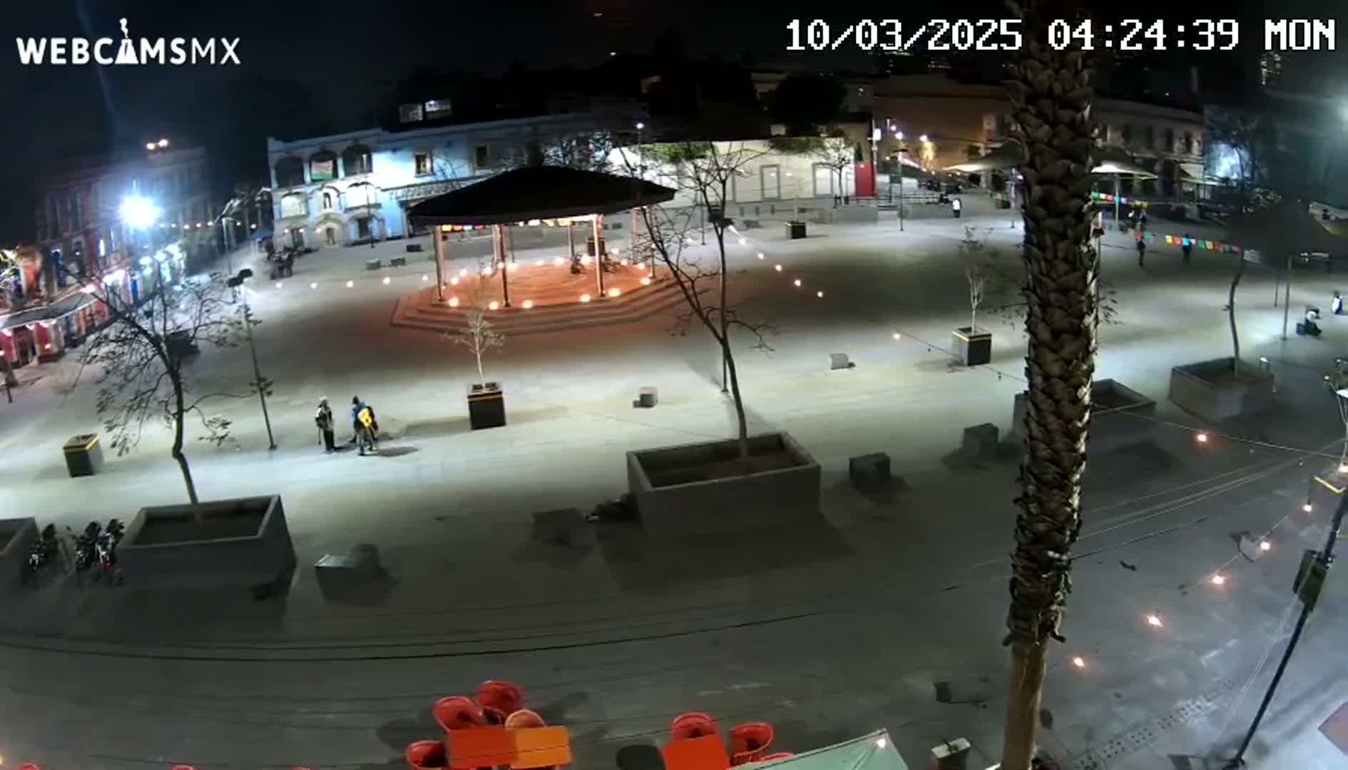 A nighttime view of a plaza with a gazebo at its center, illuminated by lights, and a few people sparsely scattered across the paved area.