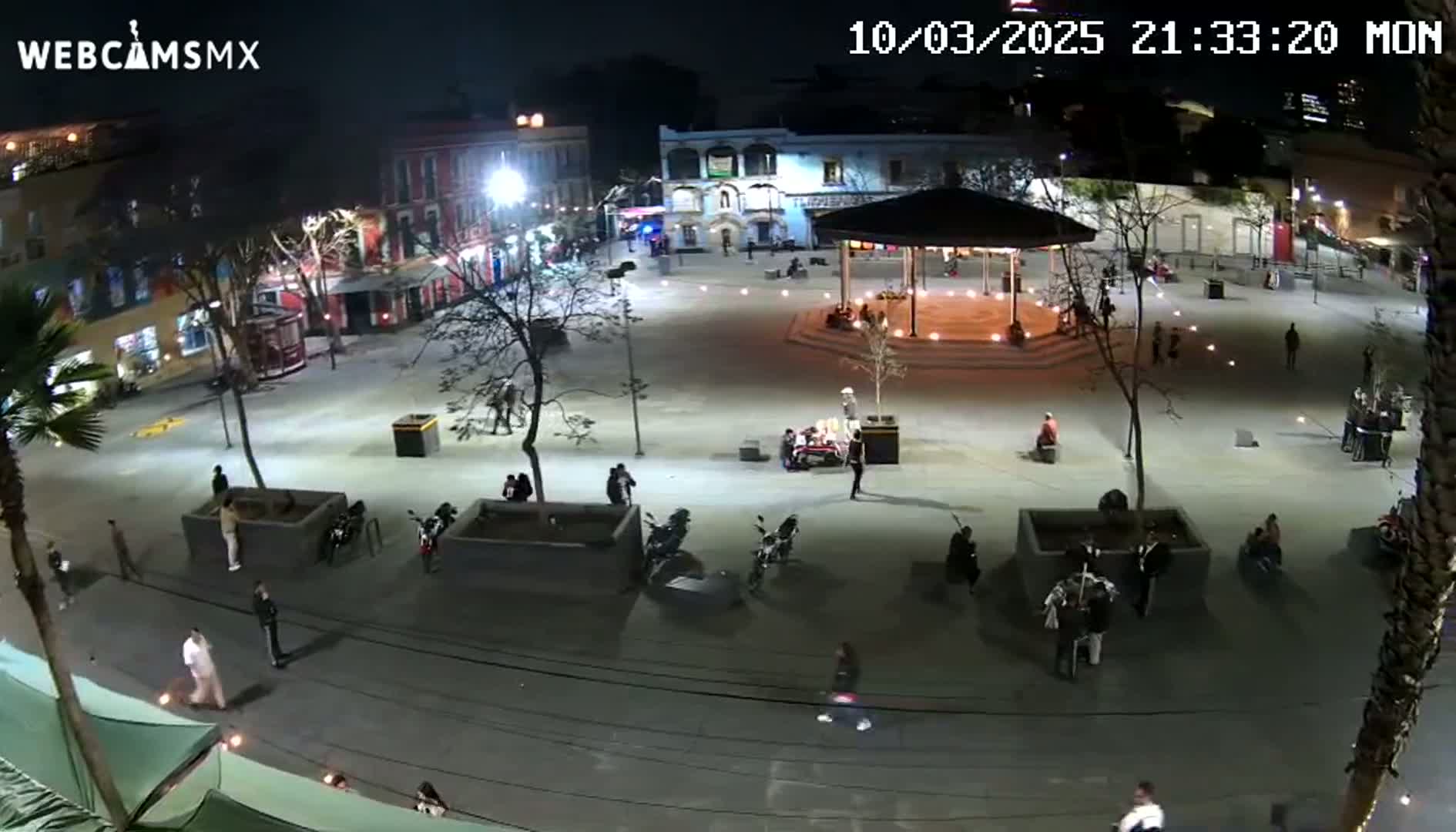 A nighttime, overhead view of a mostly empty plaza with several people scattered throughout, illuminated by streetlights and ambient lighting, featuring a gazebo in the center and surrounding buildings.