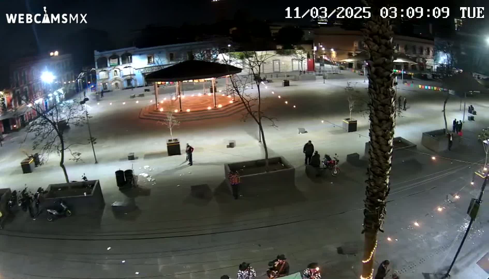 A nighttime, brightly lit plaza features a gazebo with lights, several people, and some parked motorcycles.