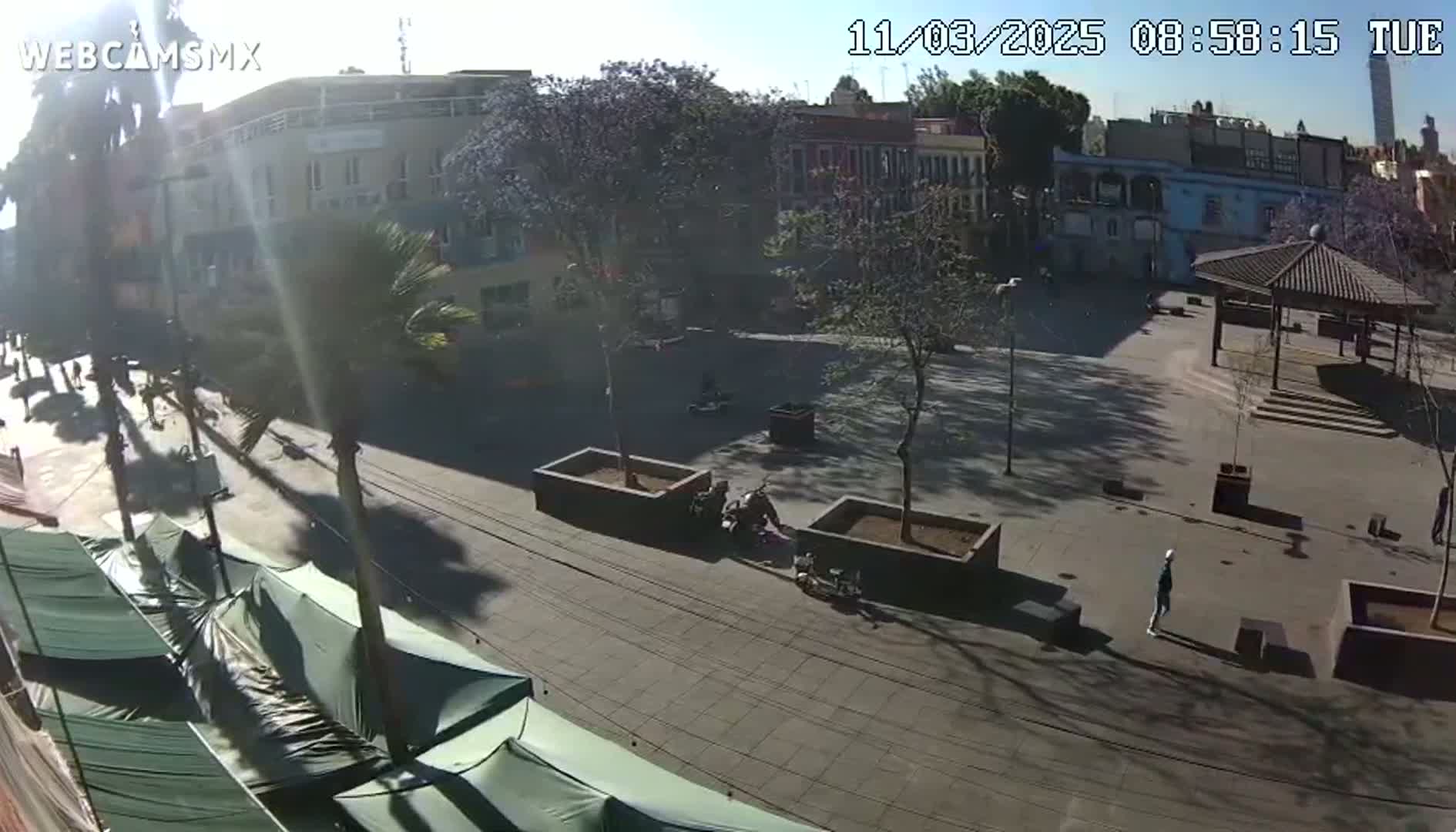 A sunny day reveals a city square with buildings, trees, and a few people; several covered market stalls are visible in the foreground.
