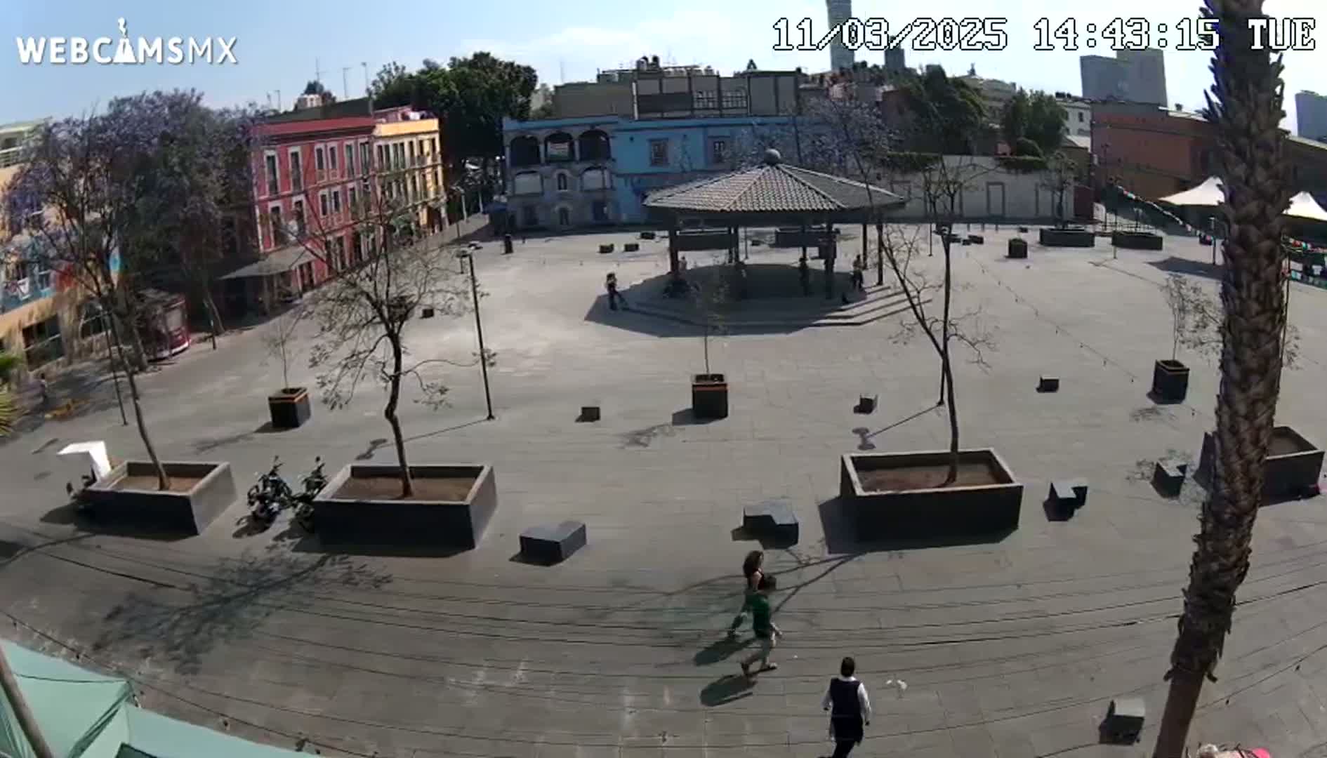 A mostly empty plaza with a gazebo in the center is surrounded by buildings under a sunny sky.
