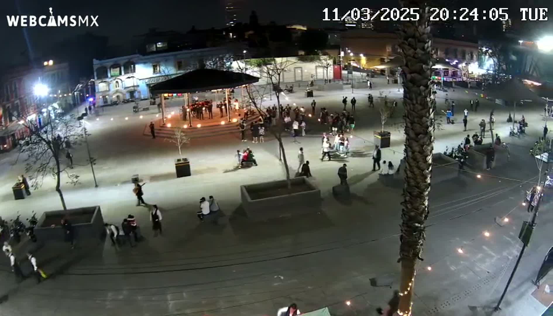 A nighttime, outdoor plaza scene shows many people gathered around a gazebo and dispersed across the paved area, illuminated by streetlights.