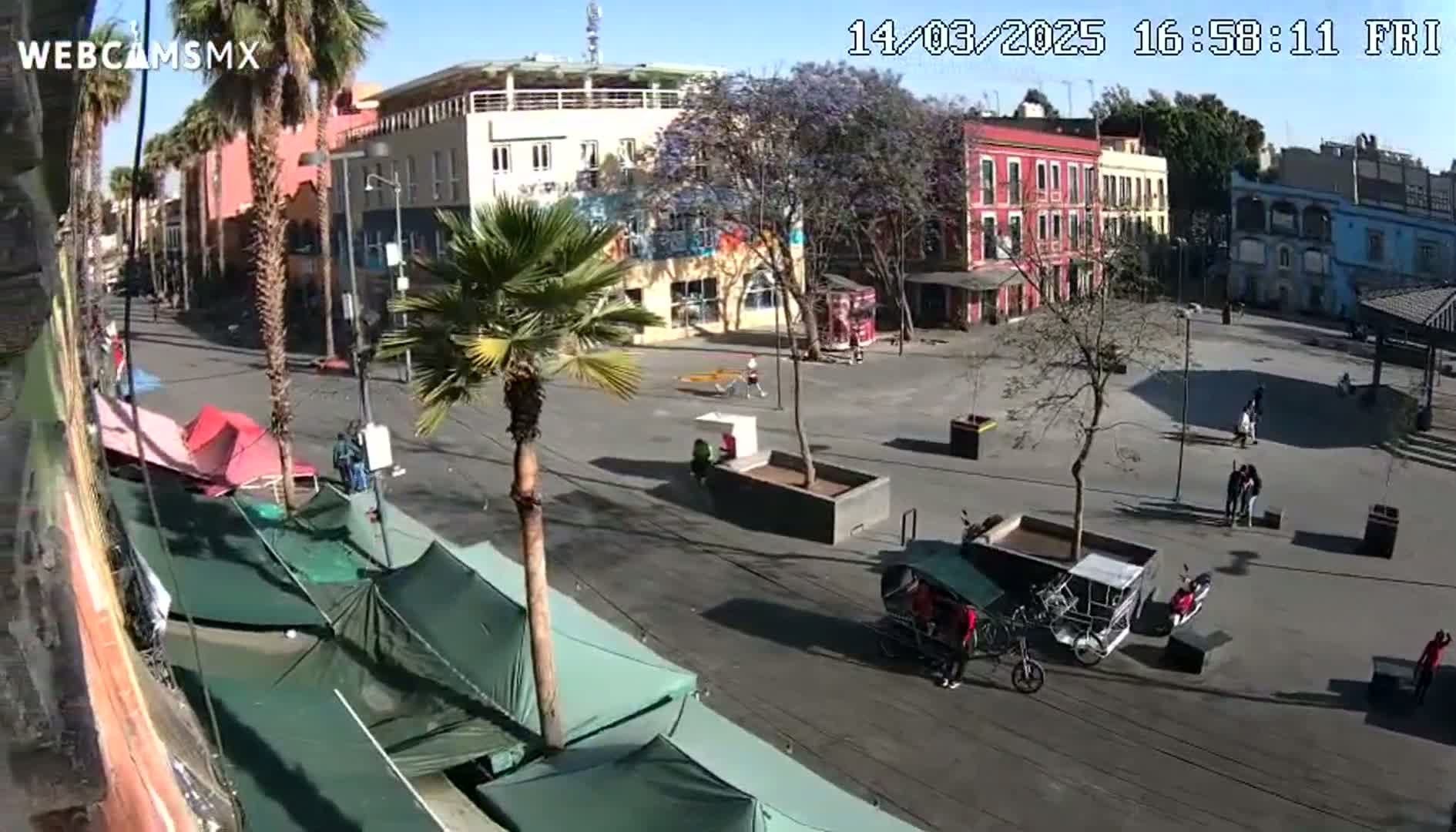 A sunny plaza with several buildings, palm trees, temporary tents, and a few people is viewed from above.
