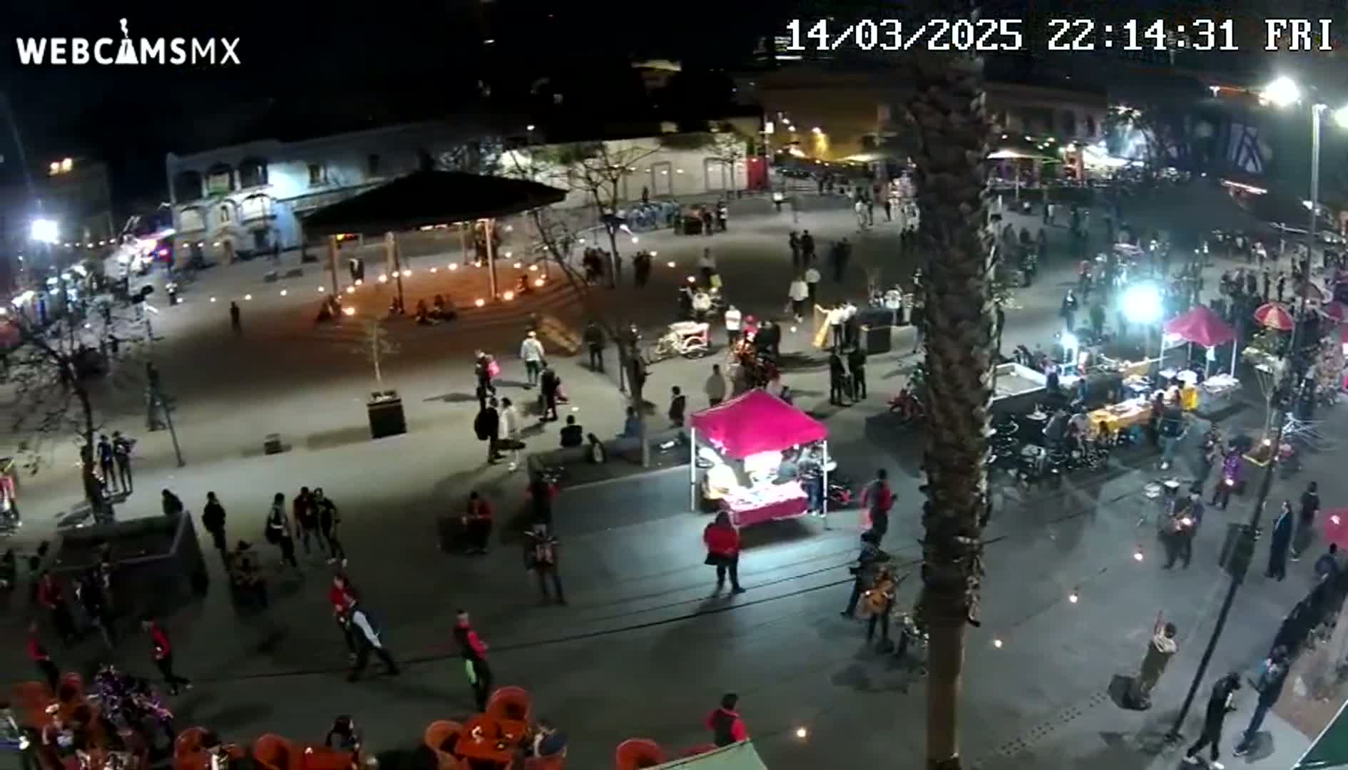 A nighttime aerial view shows a town square with numerous people, small vendors, and ambient lighting.