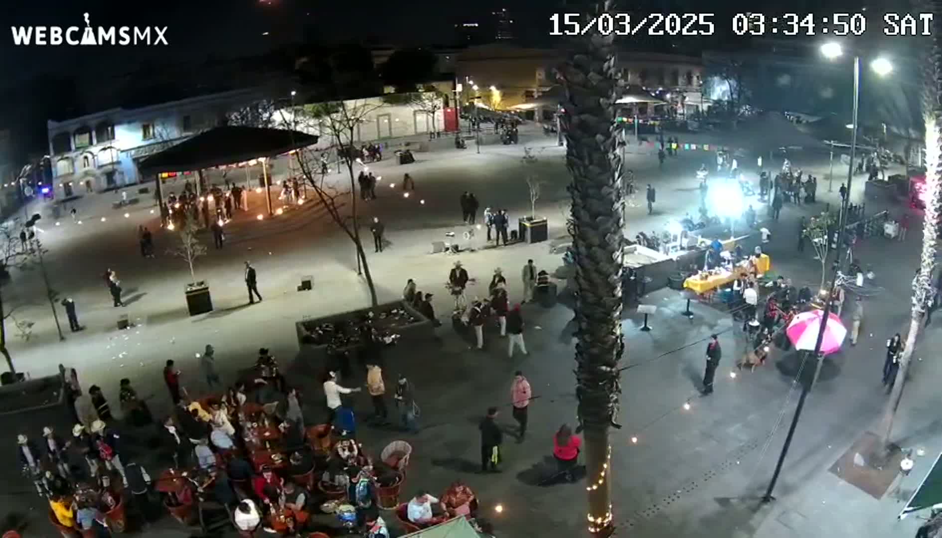 A nighttime, outdoor plaza scene shows people gathered around tables, a band playing, and various vendors or performers scattered throughout the area.