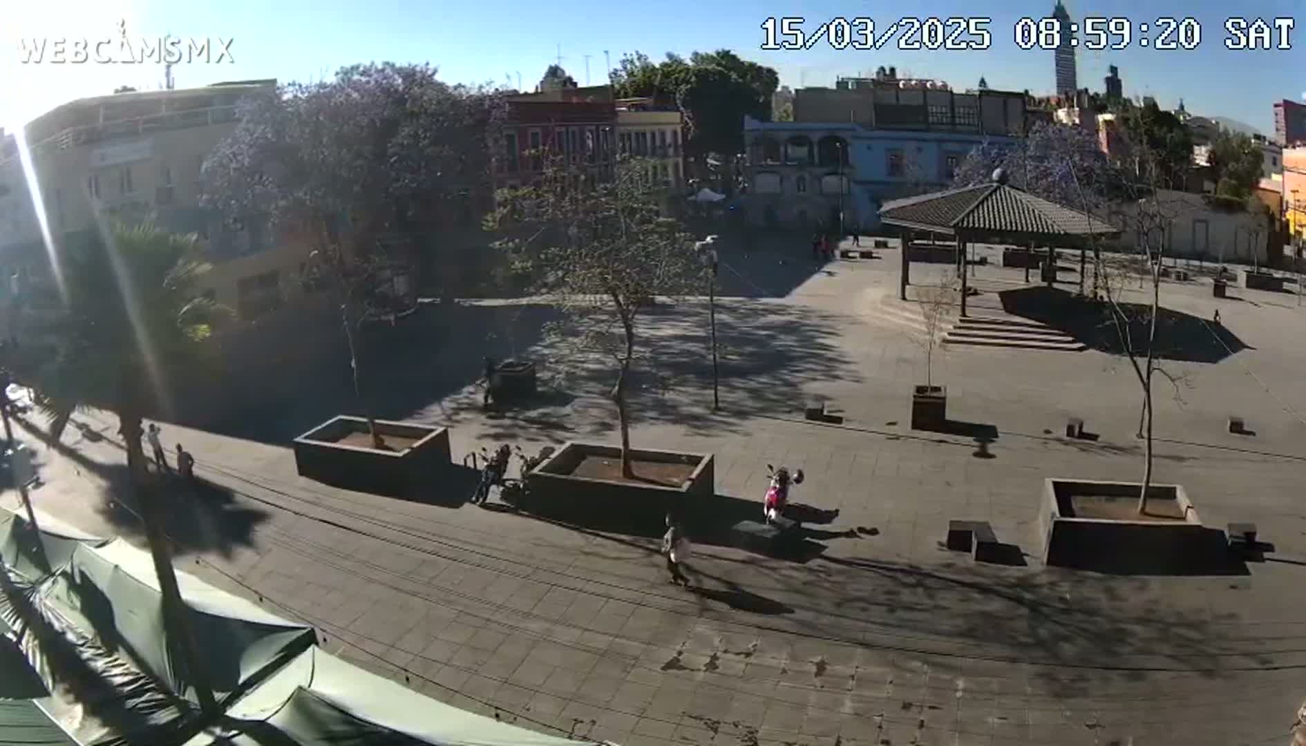 A sunny day reveals a plaza with a gazebo, several trees, and sparse pedestrians, surrounded by low buildings.