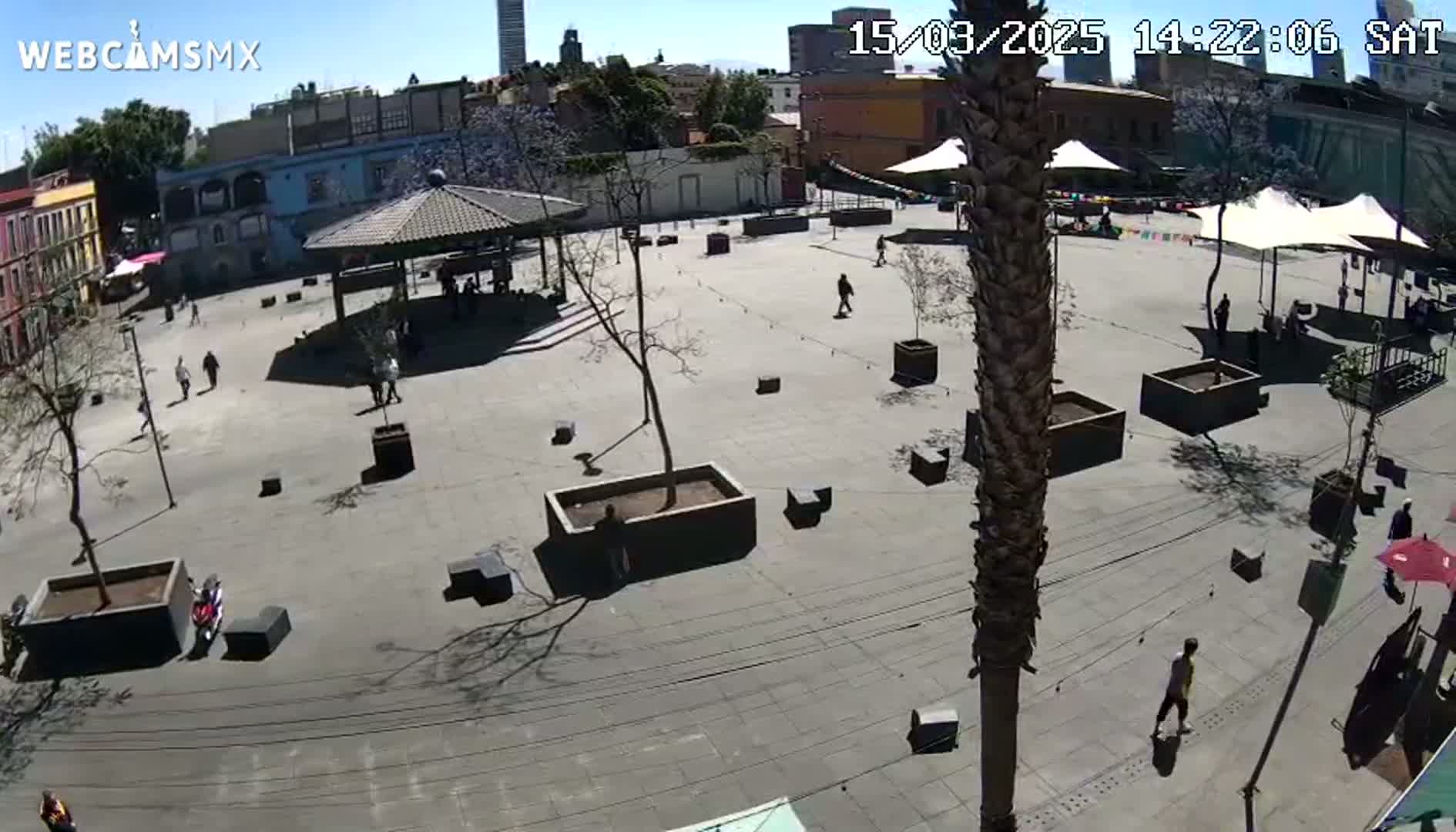 A mostly empty plaza with a few people walking around, several small rectangular planters, and a gazebo under a sunny sky.