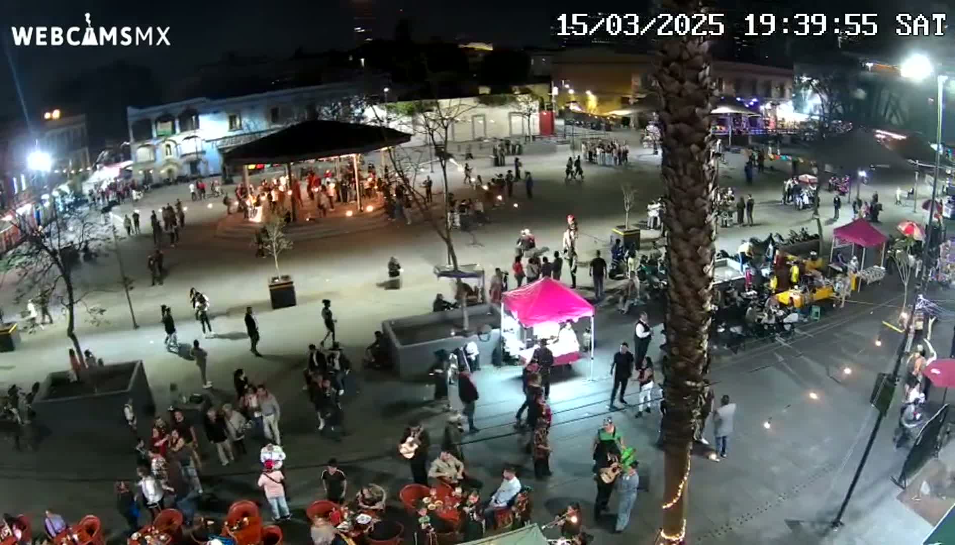 A nighttime aerial view of a town square filled with people, food stalls, and musicians, under clear skies.