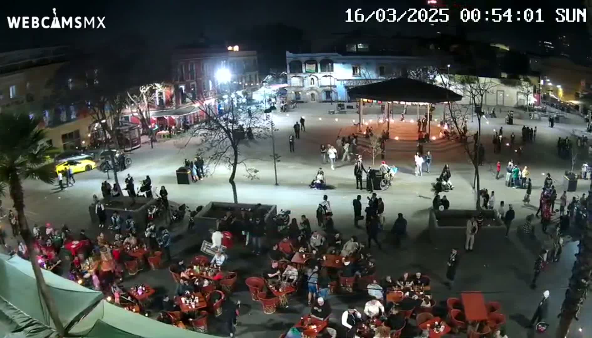 A nighttime, outdoor plaza scene shows numerous people gathered around tables and chairs at restaurants, with others walking around under the light of streetlamps.