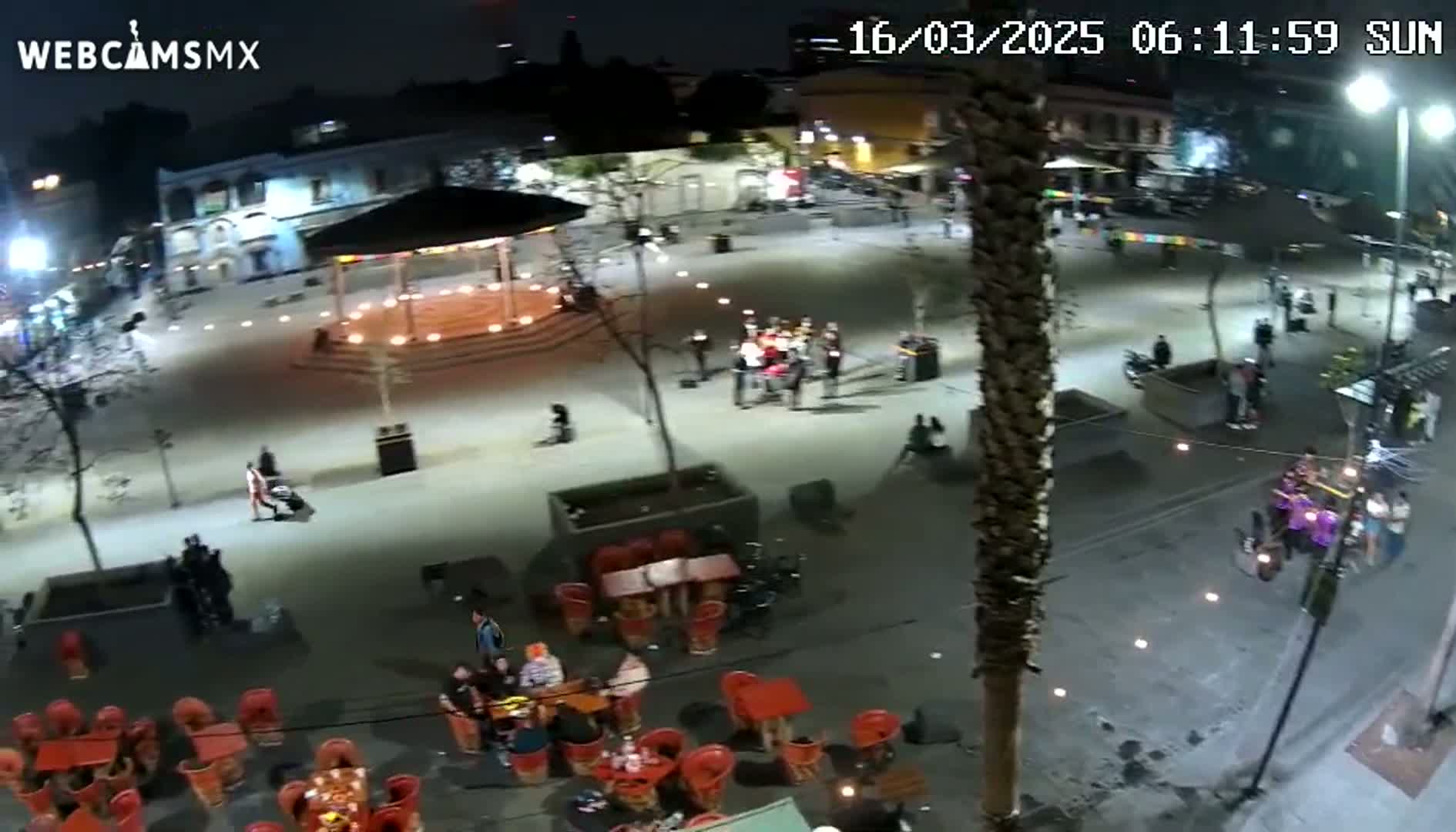 A nighttime, high-angle view shows a town square with people gathered around tables and a gazebo, under well-lit conditions.