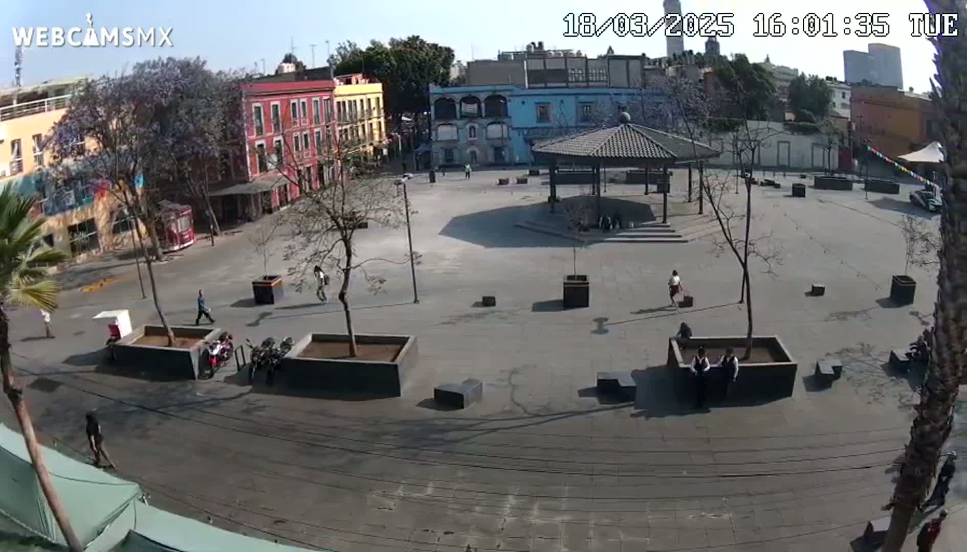 A mostly empty plaza with a gazebo in the center, surrounded by low buildings under a sunny sky.
