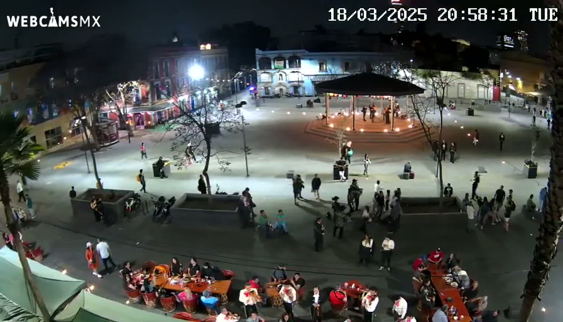 A nighttime aerial view of a town square shows many people gathered around tables, a band playing under a gazebo, and others walking around under bright lights.