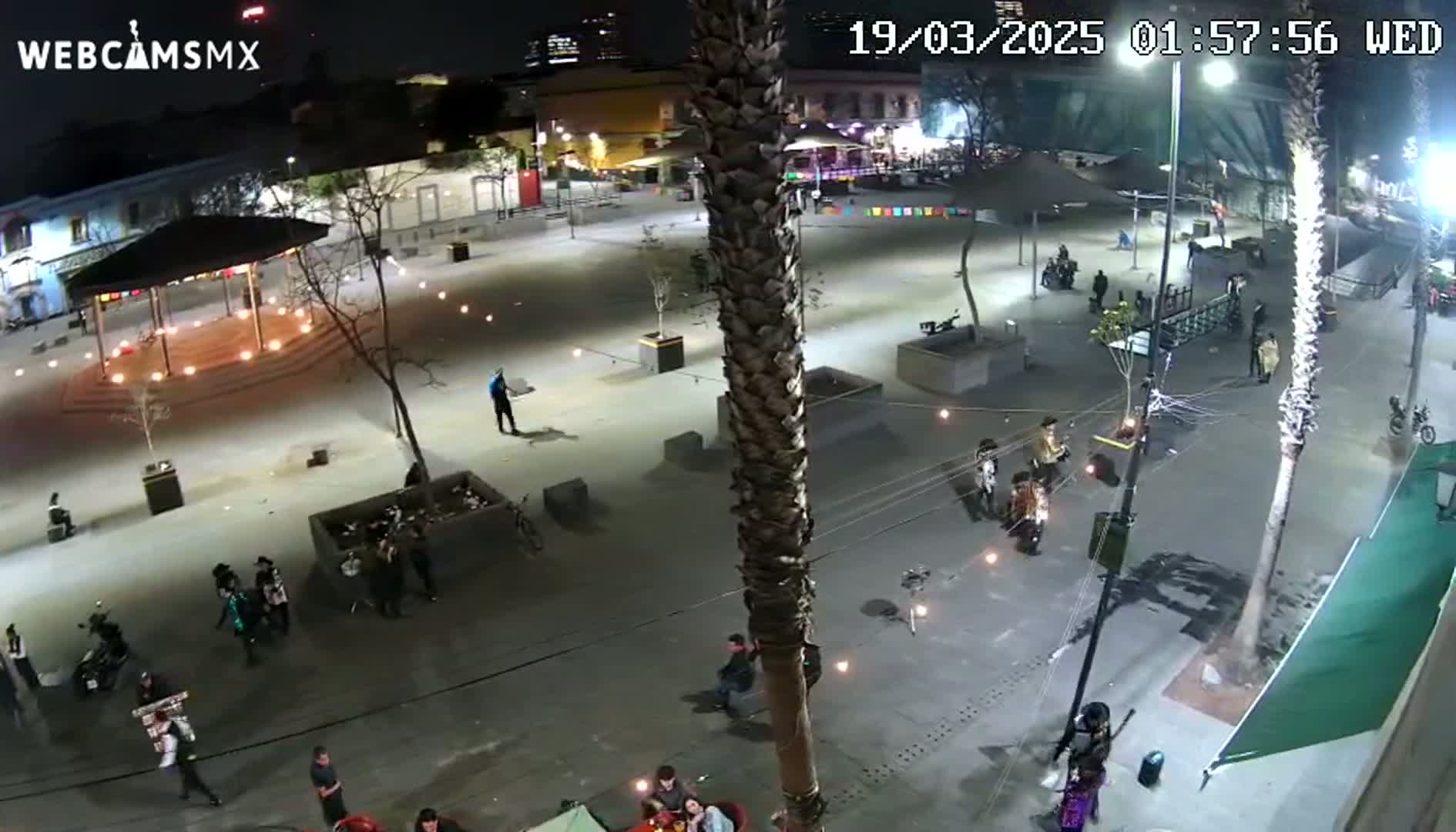 A nighttime, outdoor plaza scene shows many people gathered in small groups, with some walking around and others sitting at tables, under lights strung across the paved area, near palm trees and a gazebo.
