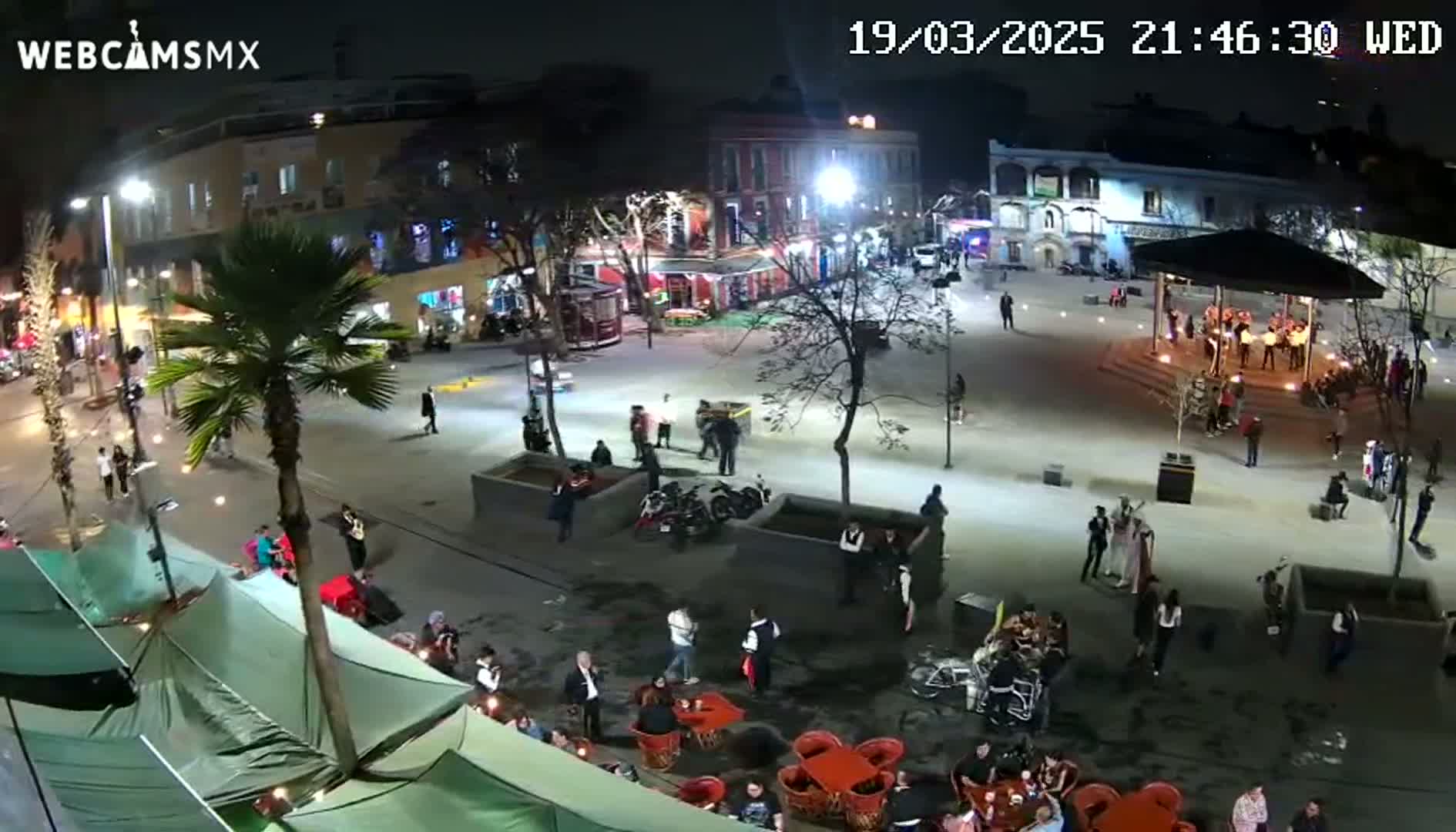 An outdoor nighttime scene shows a town square with people gathered around tables, a gazebo, and street vendors, under partly cloudy skies.