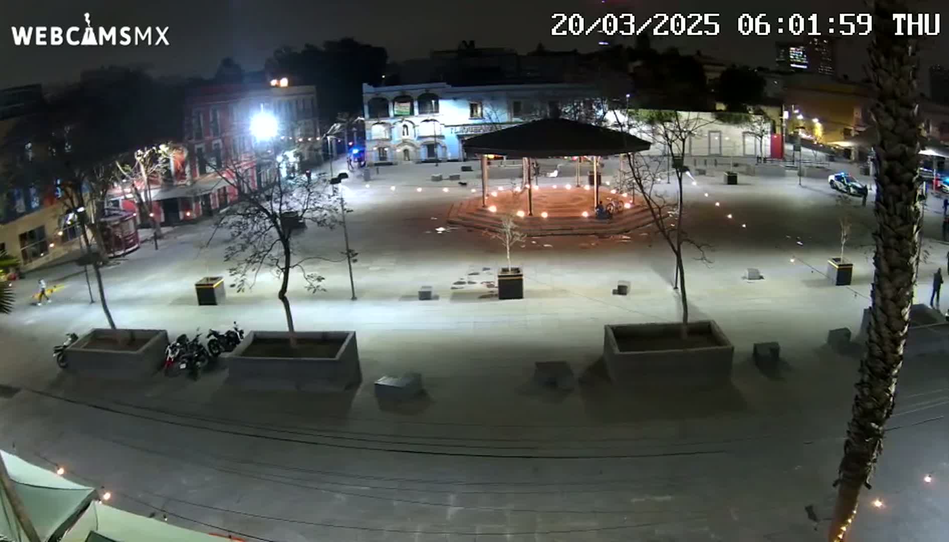 A nighttime, brightly lit plaza with a gazebo at its center, surrounded by buildings and trees, and several parked motorcycles.