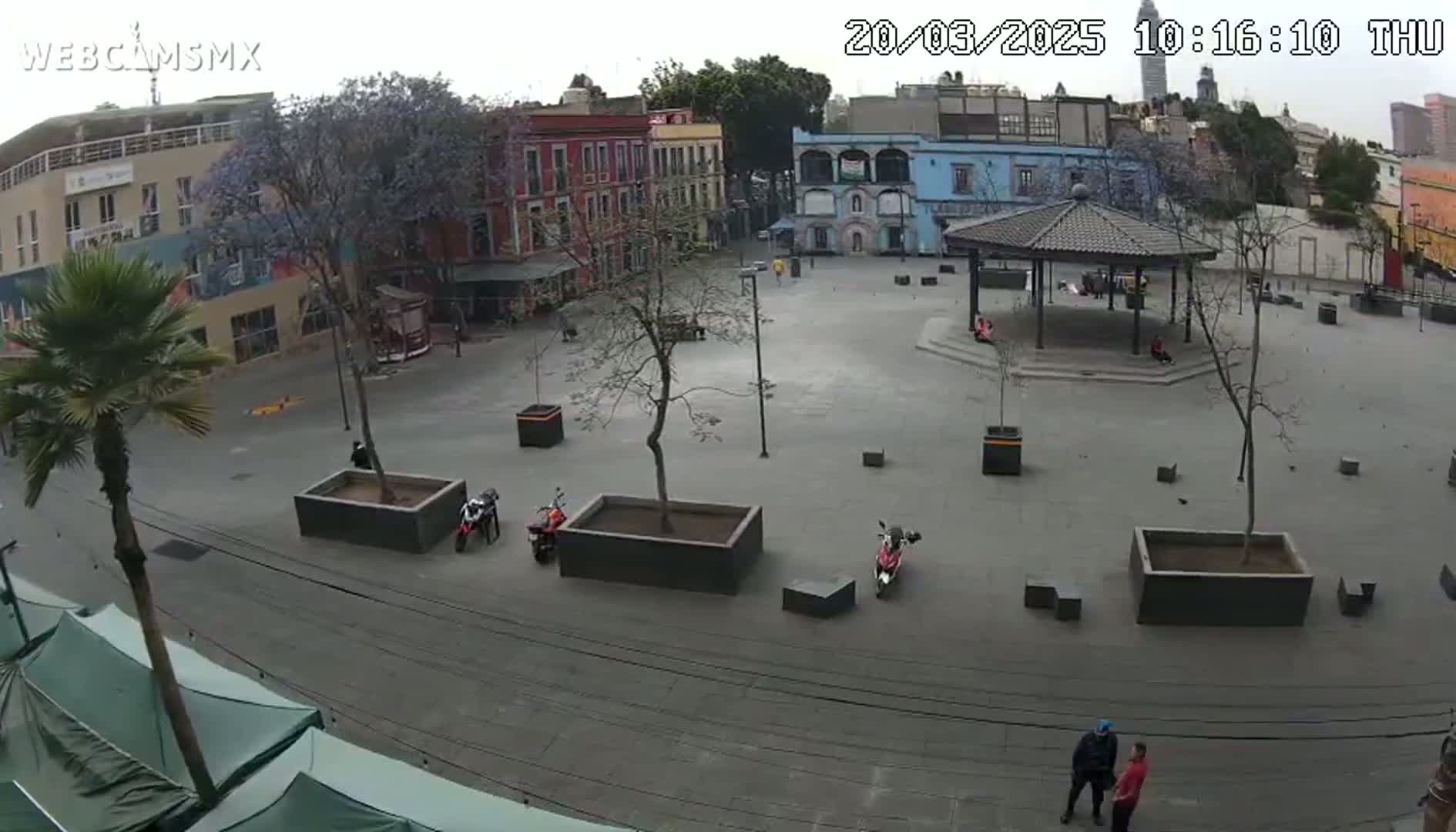 A mostly empty plaza with several parked motorcycles, a gazebo, and a few people is seen on an overcast day.