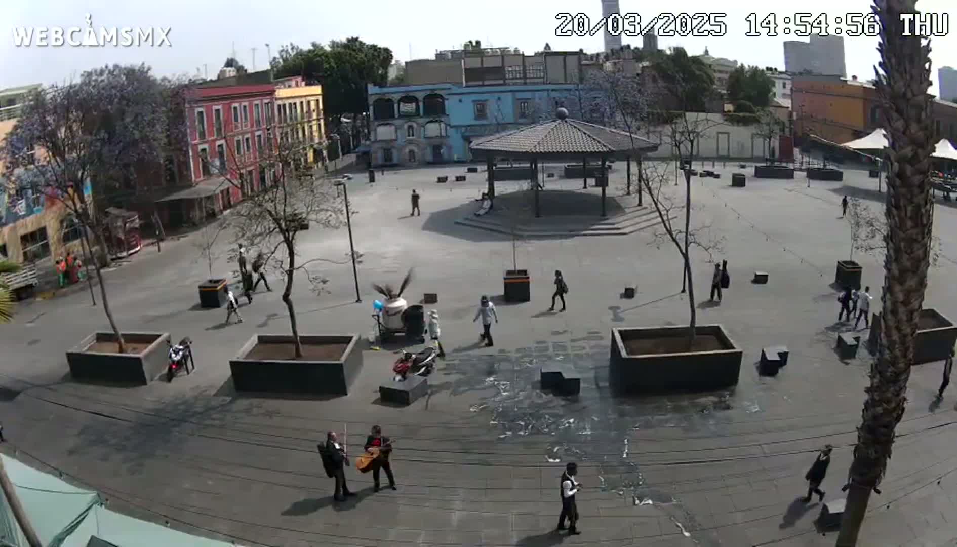 A mostly empty plaza with a gazebo, surrounded by colorful buildings, under a partly sunny sky, with several people walking around and two musicians playing string instruments.