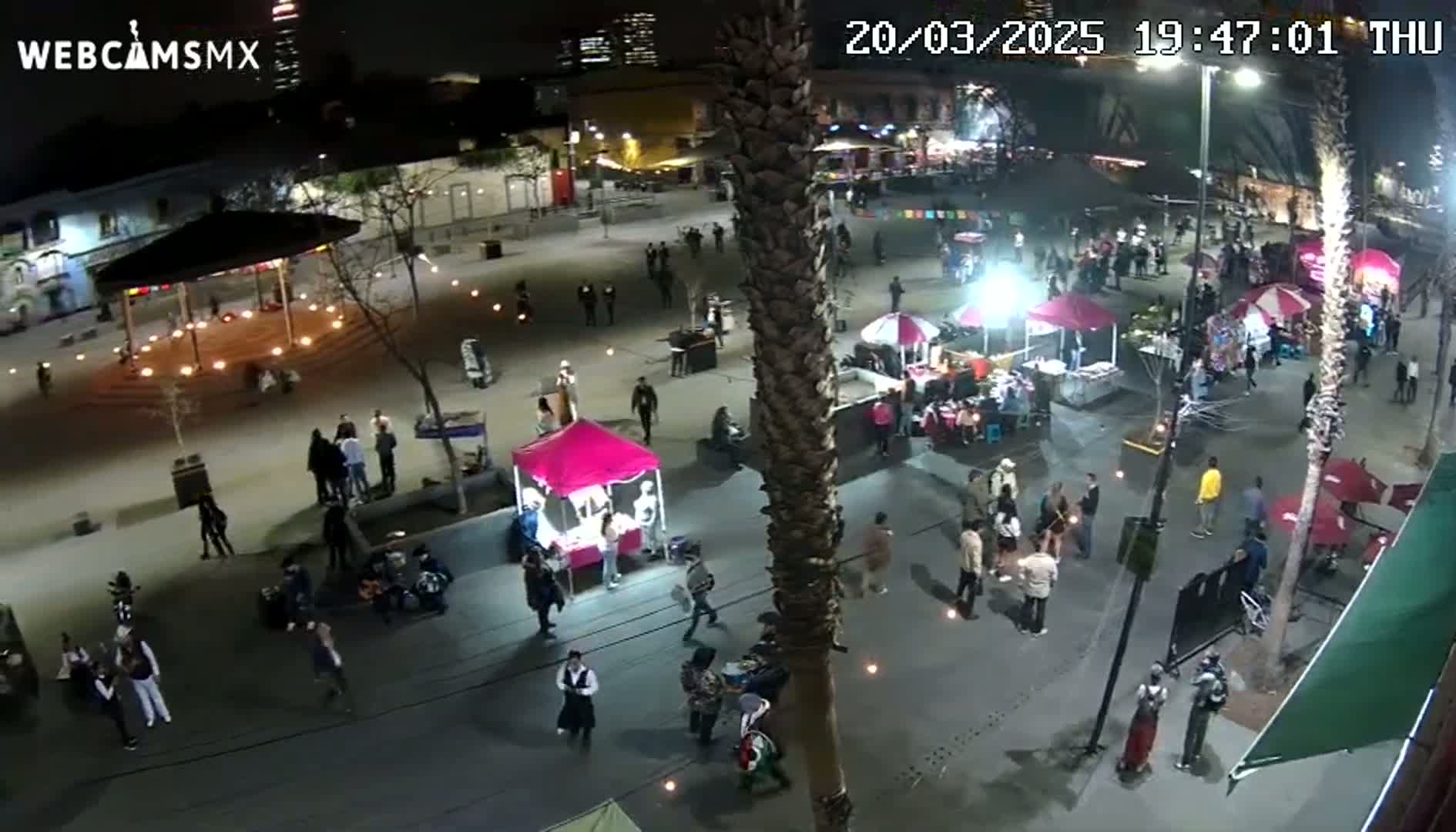 A nighttime aerial view of a plaza filled with people, market stalls, and a gazebo, under clear skies.
