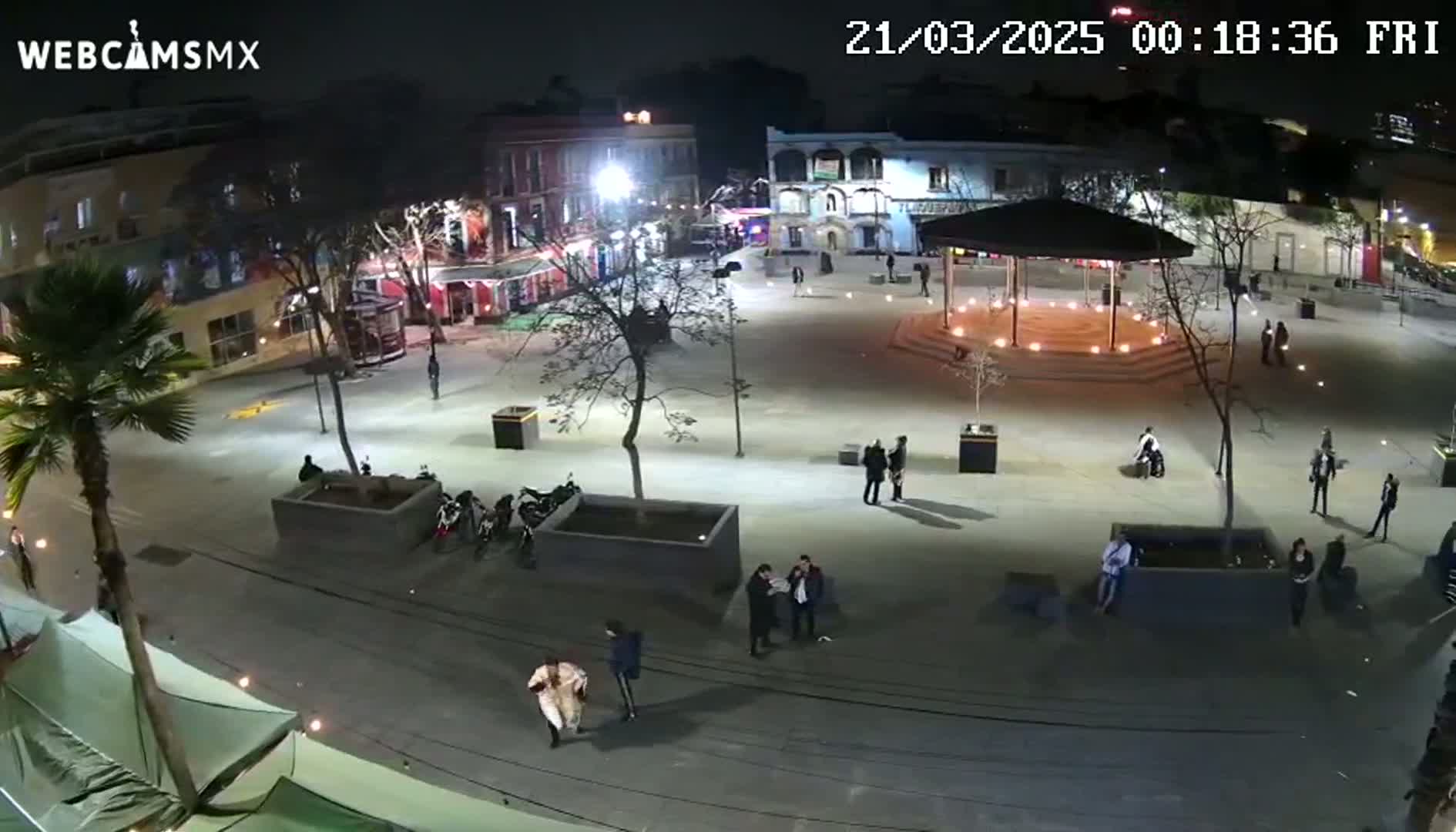 A nighttime aerial view of a mostly empty town square with a gazebo, several people, and parked motorcycles under artificial light.