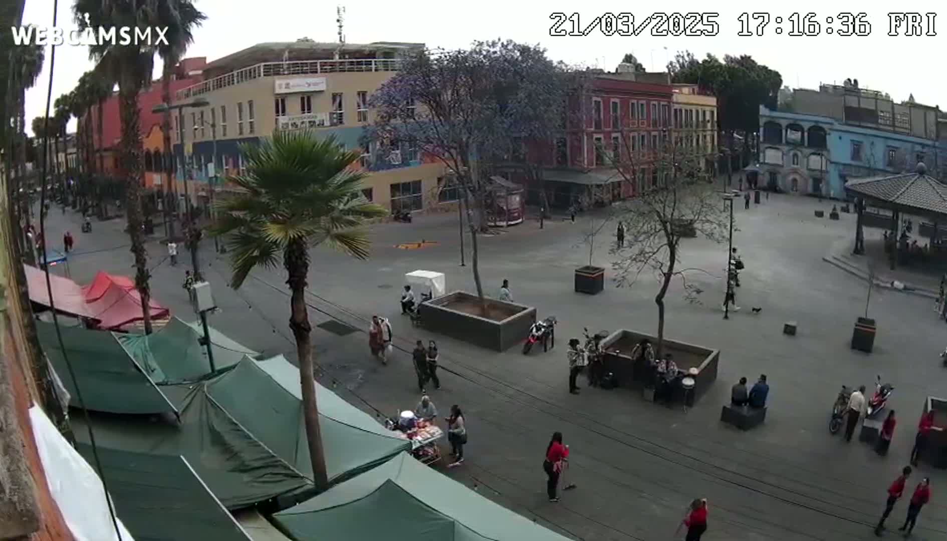 A mostly empty town square with several people and market stalls is viewed from above on an overcast day.