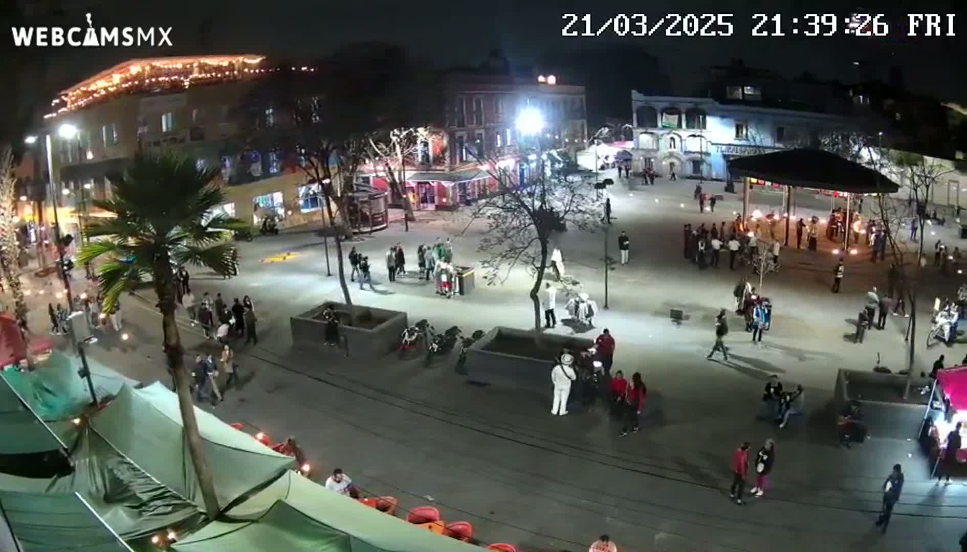 A nighttime aerial view of a town square with many people gathered around a gazebo and food stalls under clear skies.