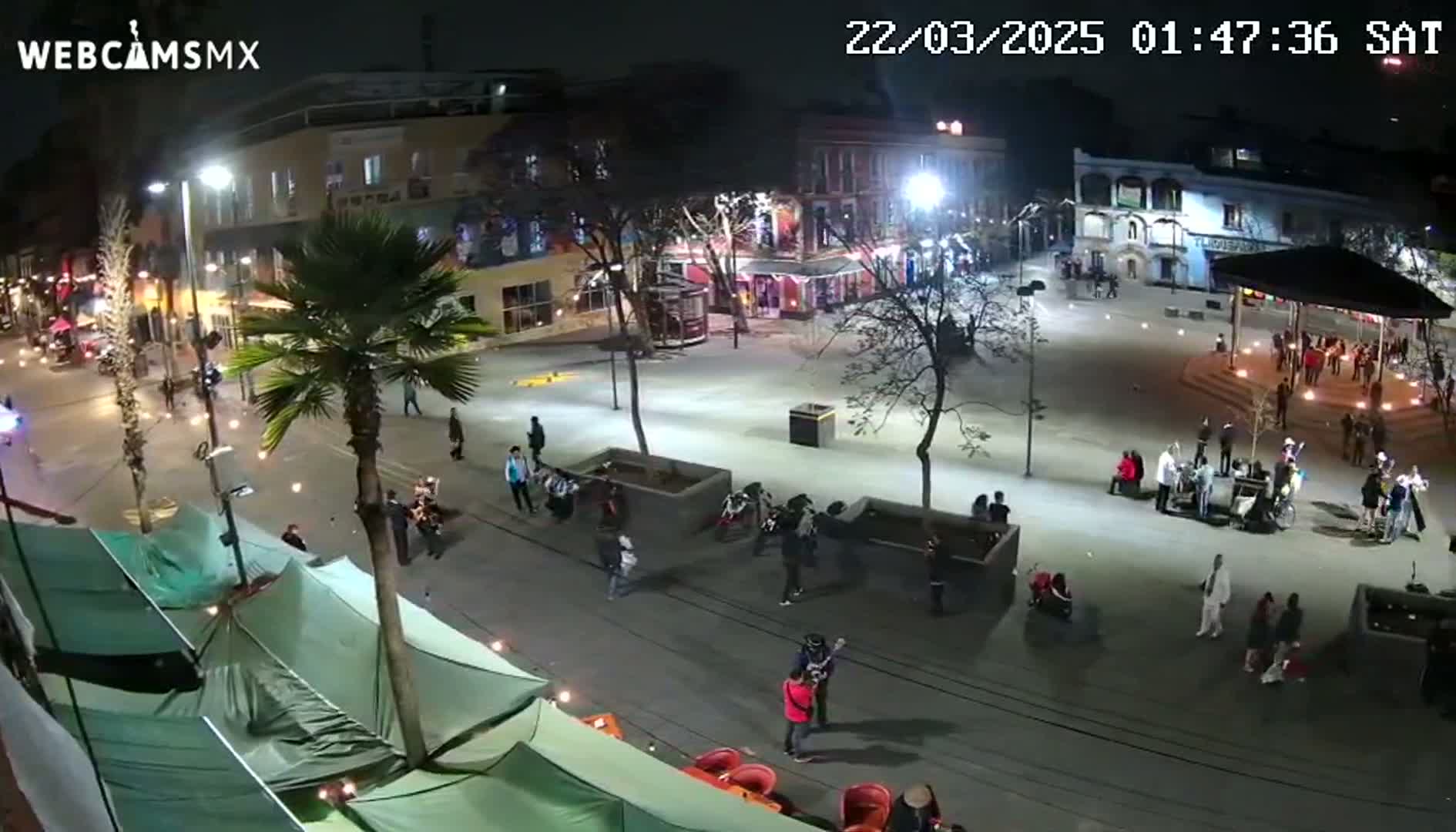 A nighttime aerial view of a city square with people gathered, some standing in groups and others walking around, under illuminated streetlights.