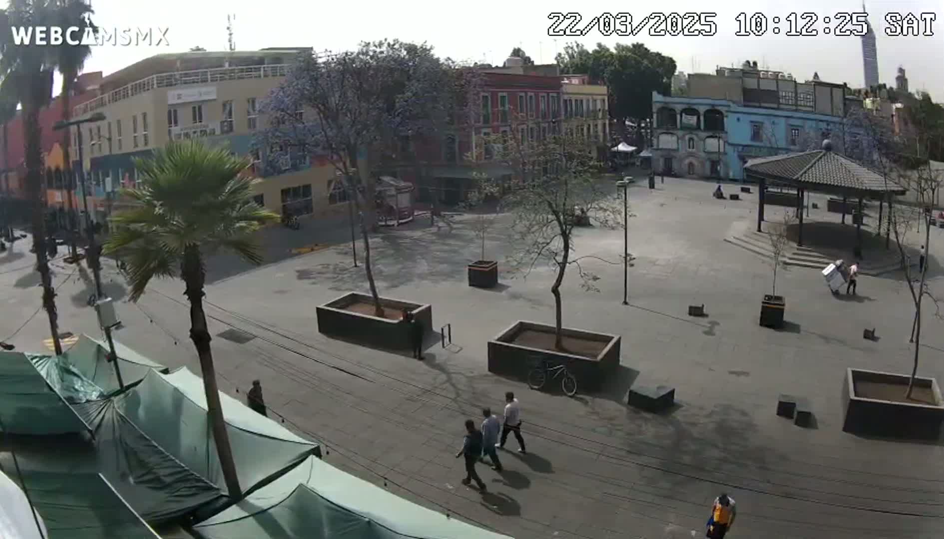 A mostly empty plaza with a few pedestrians, some buildings, and several palm trees is visible under a bright, sunny sky.