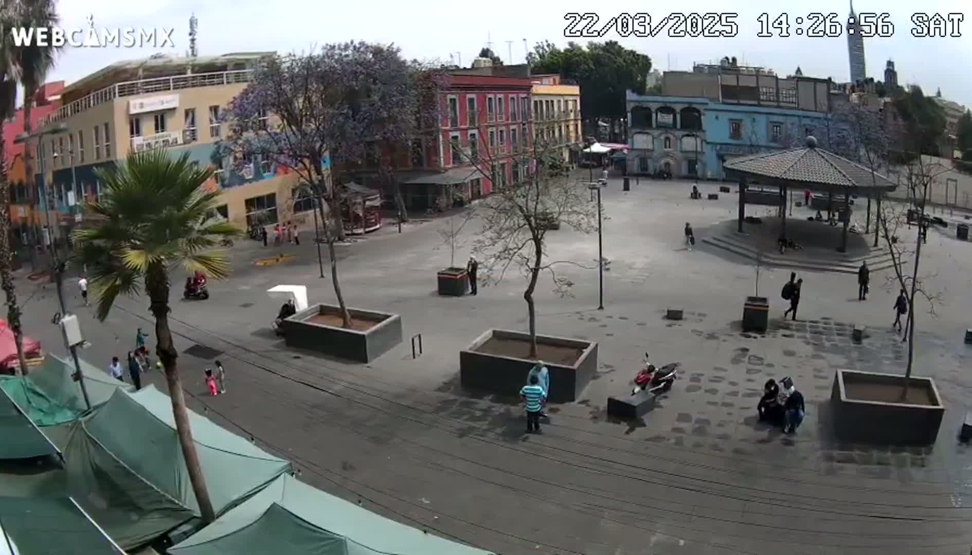 A mostly empty plaza with several buildings surrounding it, some trees, and a few people are visible on a partly sunny day.